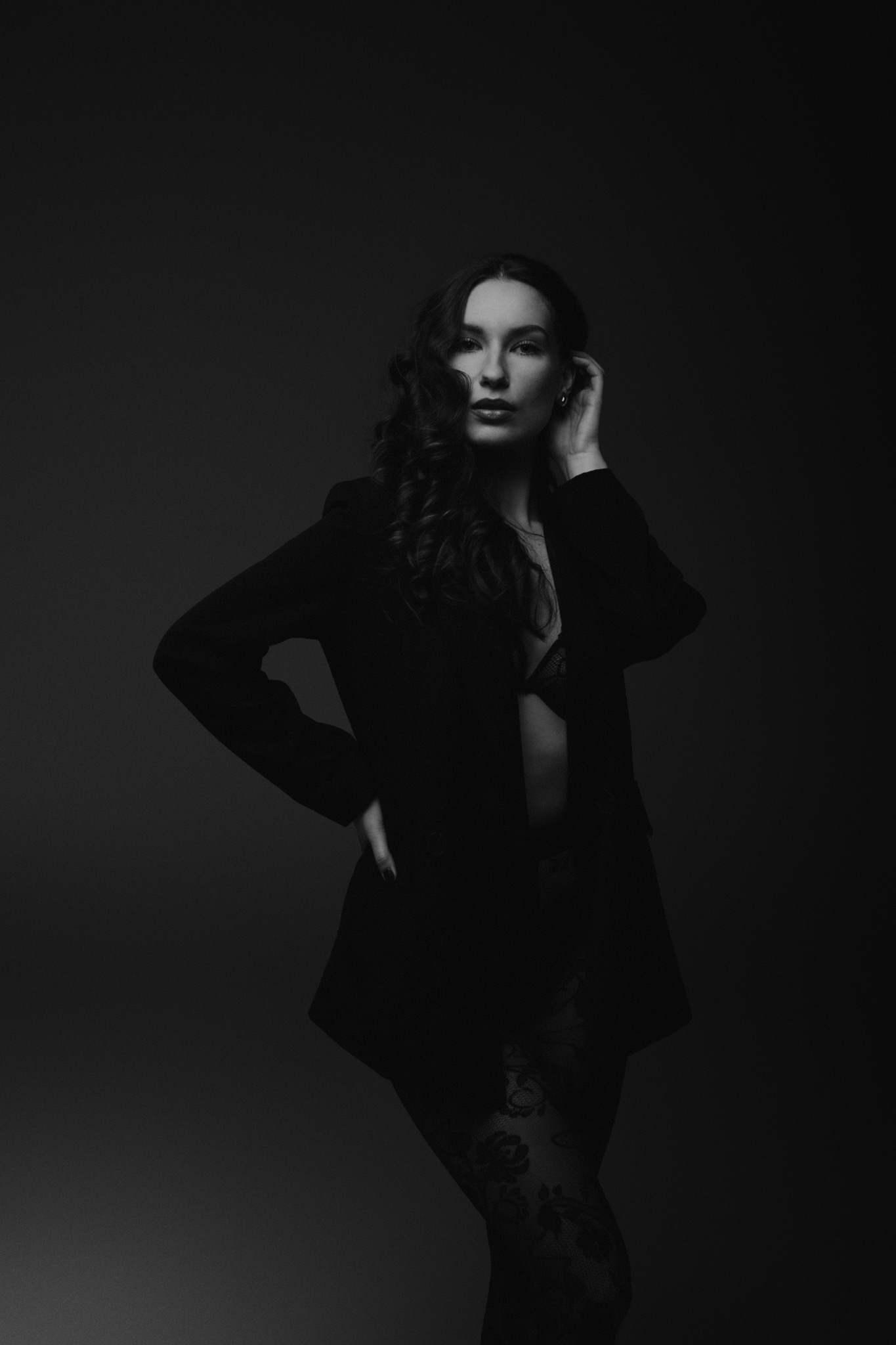 Portret kobiety w czarnej marynarce i koronkowej bieliźnie, pozującej na ciemnym tle w studiu, z ręką na biodrze i drugą dotykającą włosów.