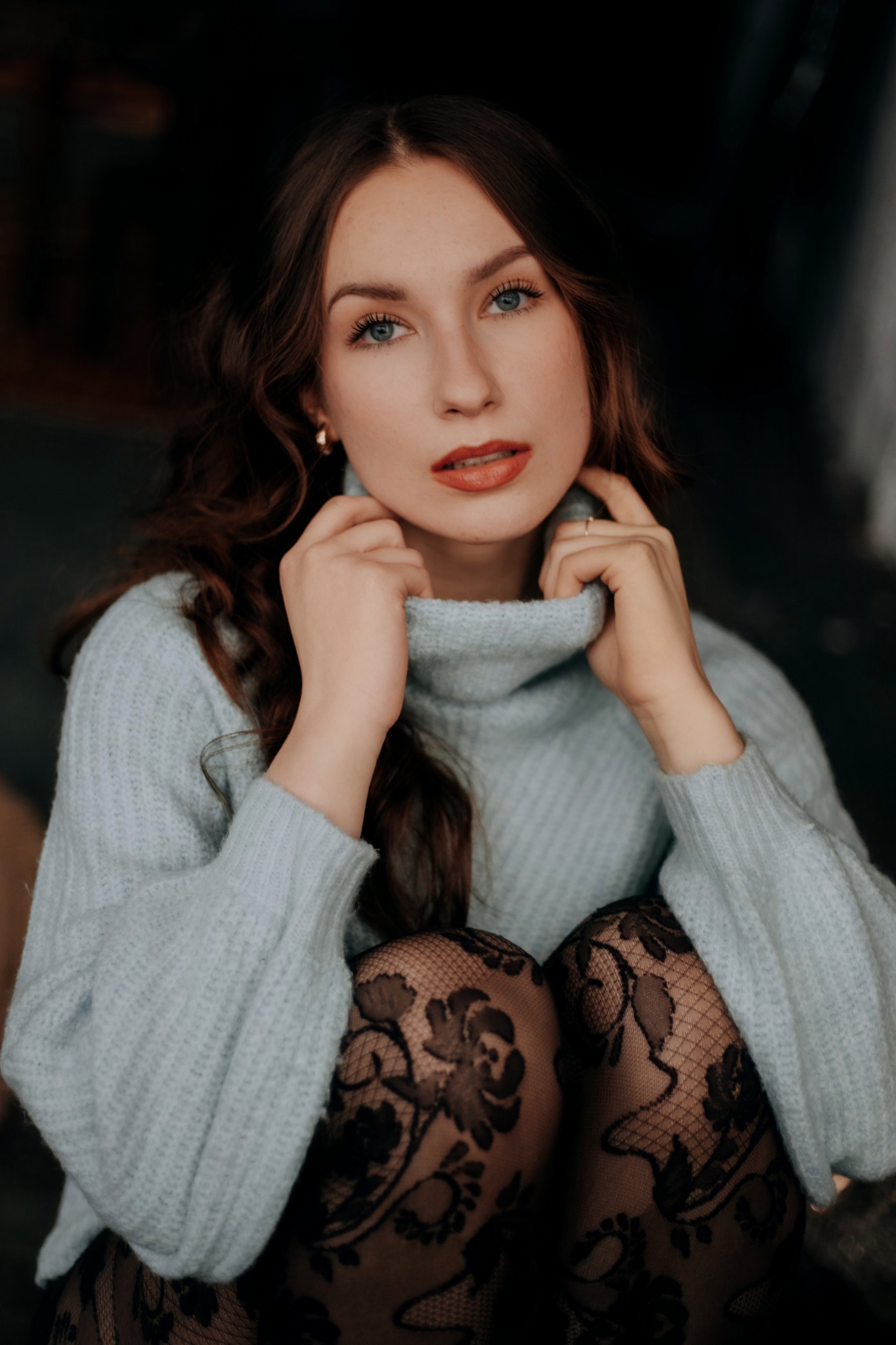 Portret kobiety o jasnych oczach w niebieskim swetrze z golfem i czarnych rajstopach w kwiatowe wzory, delikatny makijaż, złote kolczyki.