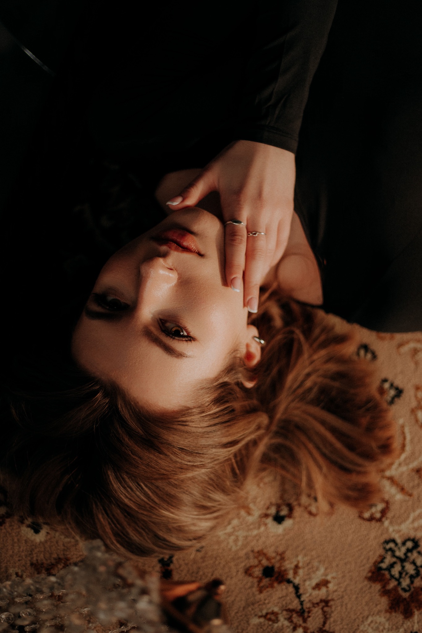 Portret kobiety leżącej na wzorzystym dywanie, z dłonią przy twarzy, pierścionkami na palcach i delikatnym makijażem w ciepłym świetle.