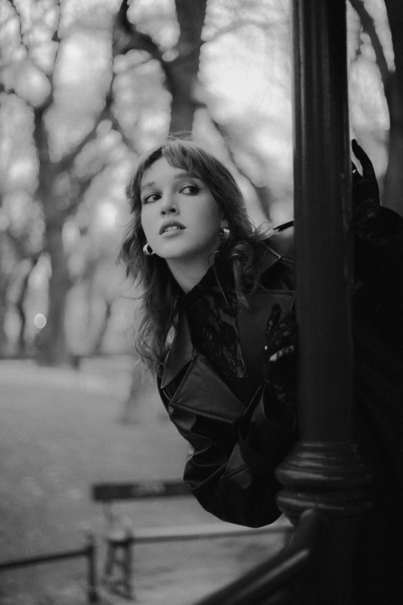 Czarno-białe ujęcie kobiety w skórzanym płaszczu, spoglądającej w bok zza latarni w krakowskim parku, z delikatnym uśmiechem na twarzy.