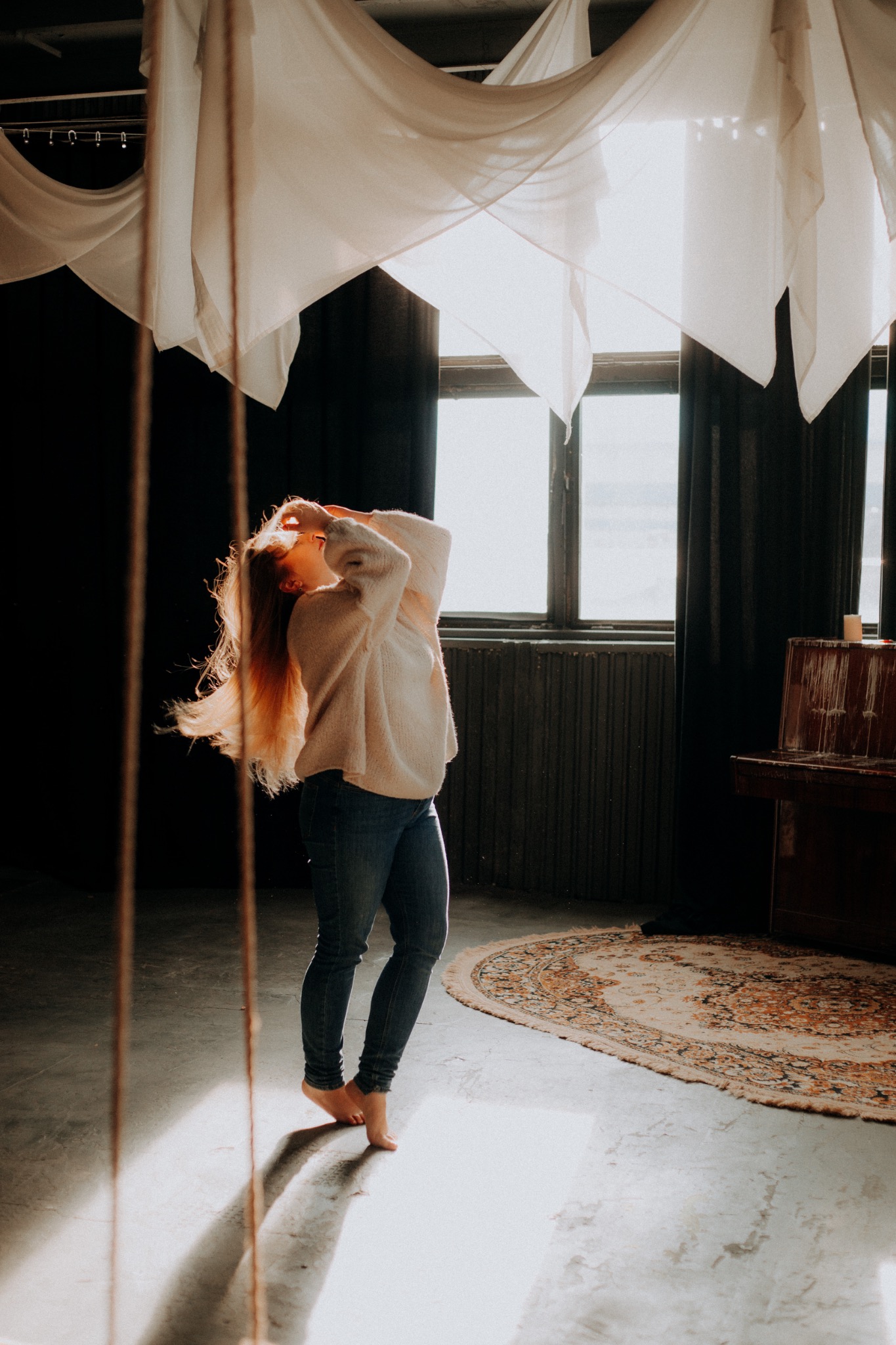 Kobieta w swetrze i jeansach, stojąca boso na tle okna i dekoracyjnych tkanin, z rozpuszczonymi, rudymi włosami w ruchu, oświetlona ciepłym światłem.