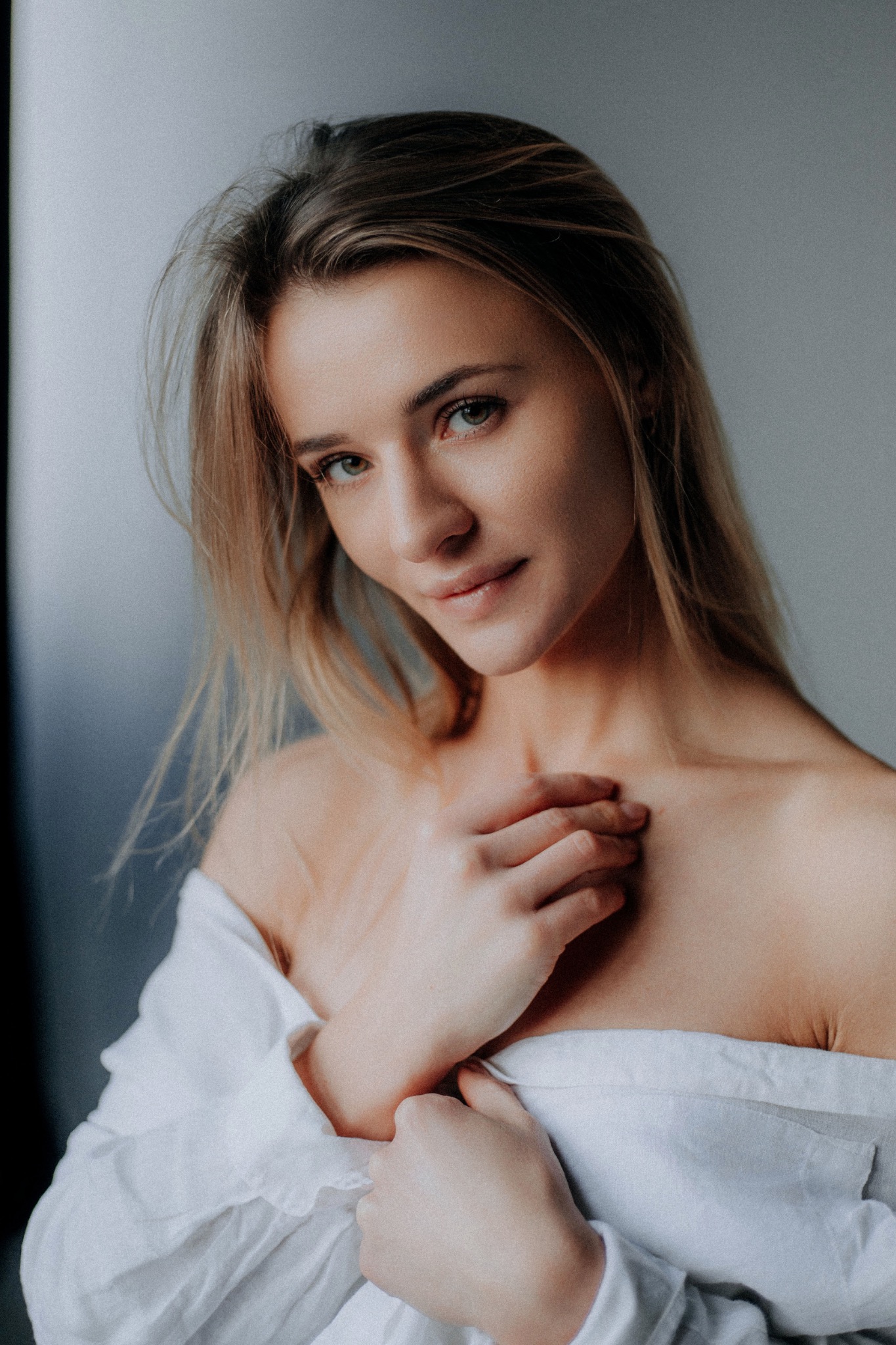 Portret kobiety w białej koszuli, delikatne światło, subtelny uśmiech, dłonie na dekolcie, naturalny makijaż, rozpuszczone blond włosy.