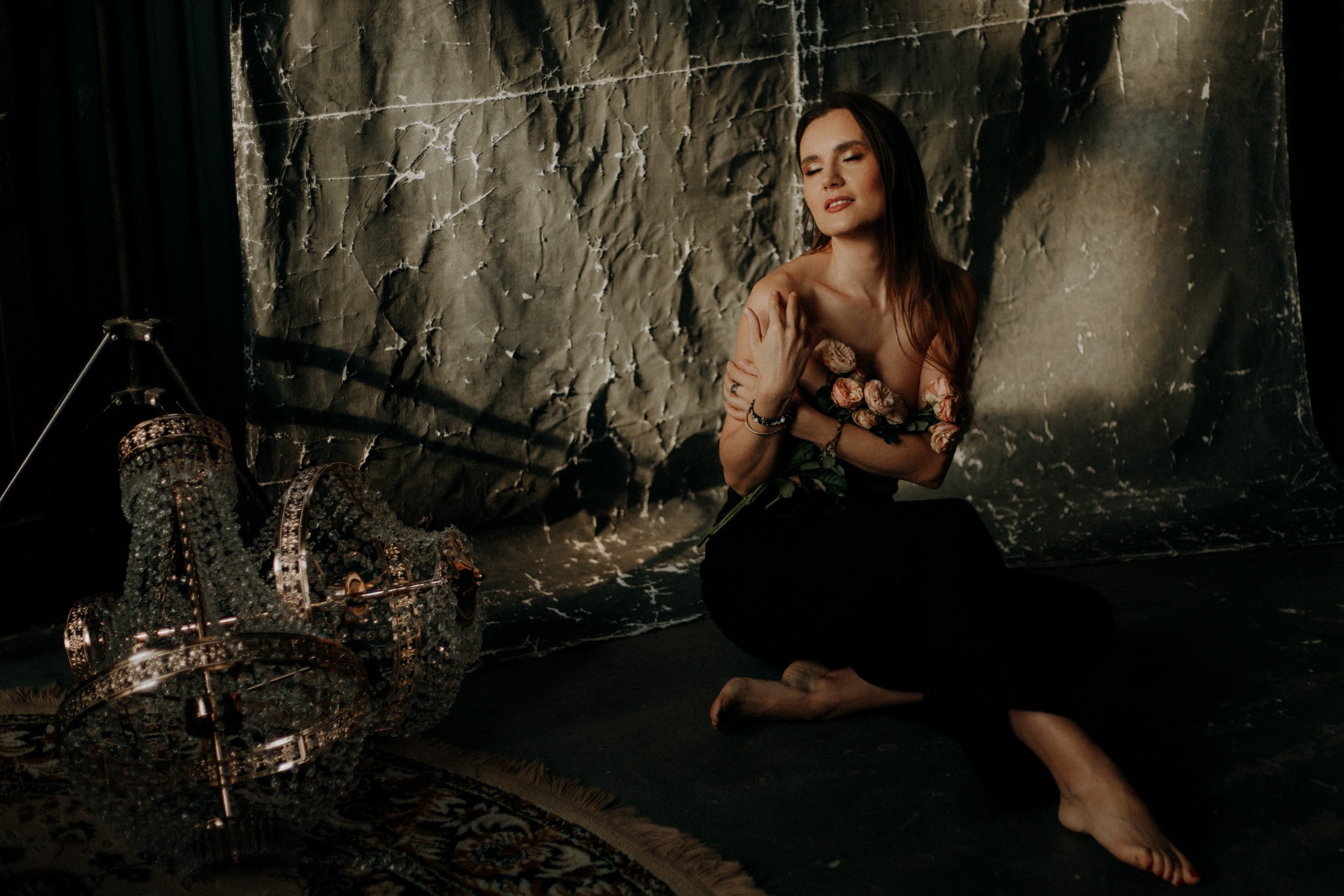 Kobieta siedząca na podłodze z bukietem różowych róż, w tle teksturowana ściana i kryształowy żyrandol