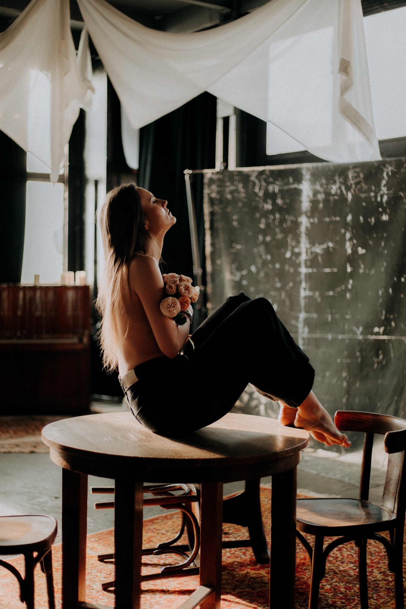 Kobieta siedząca na stole, trzymająca bukiet różowych kwiatów, z głową zadartą do góry, w tle okno i draperie.