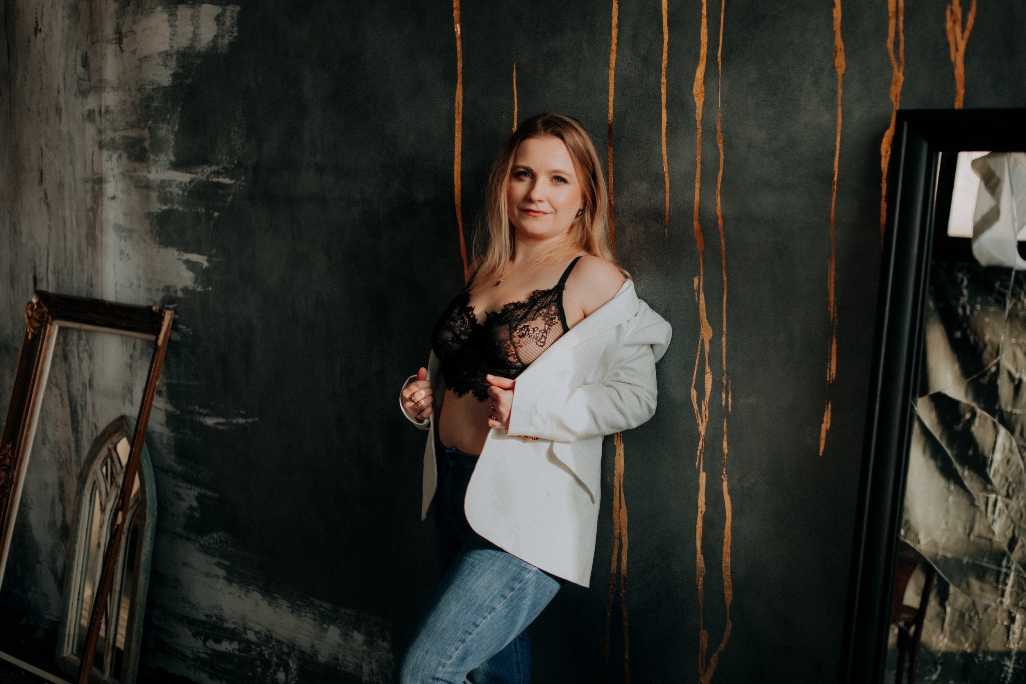 Kobieta w koronkowej bieliźnie i białej marynarce pozuje na tle ściany w odcieniach szarości z złotymi akcentami, obok widoczne ramy obrazów.