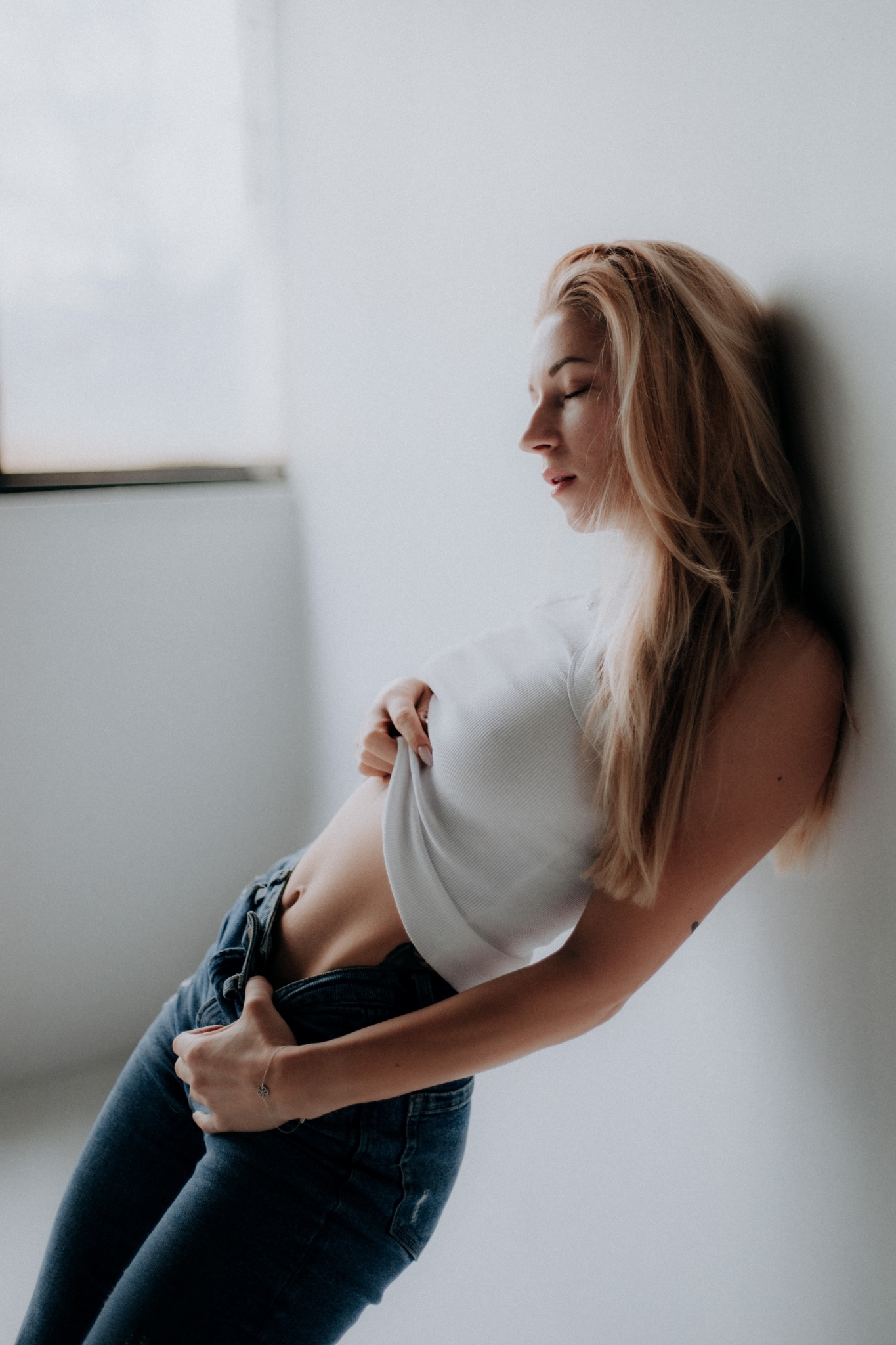 Kobieta o blond włosach, oparta o białą ścianę, w białym topie i dżinsach, z niedopiętymi spodniami, delikatny profil twarzy, subtelna bransoletka na nadgarstku.