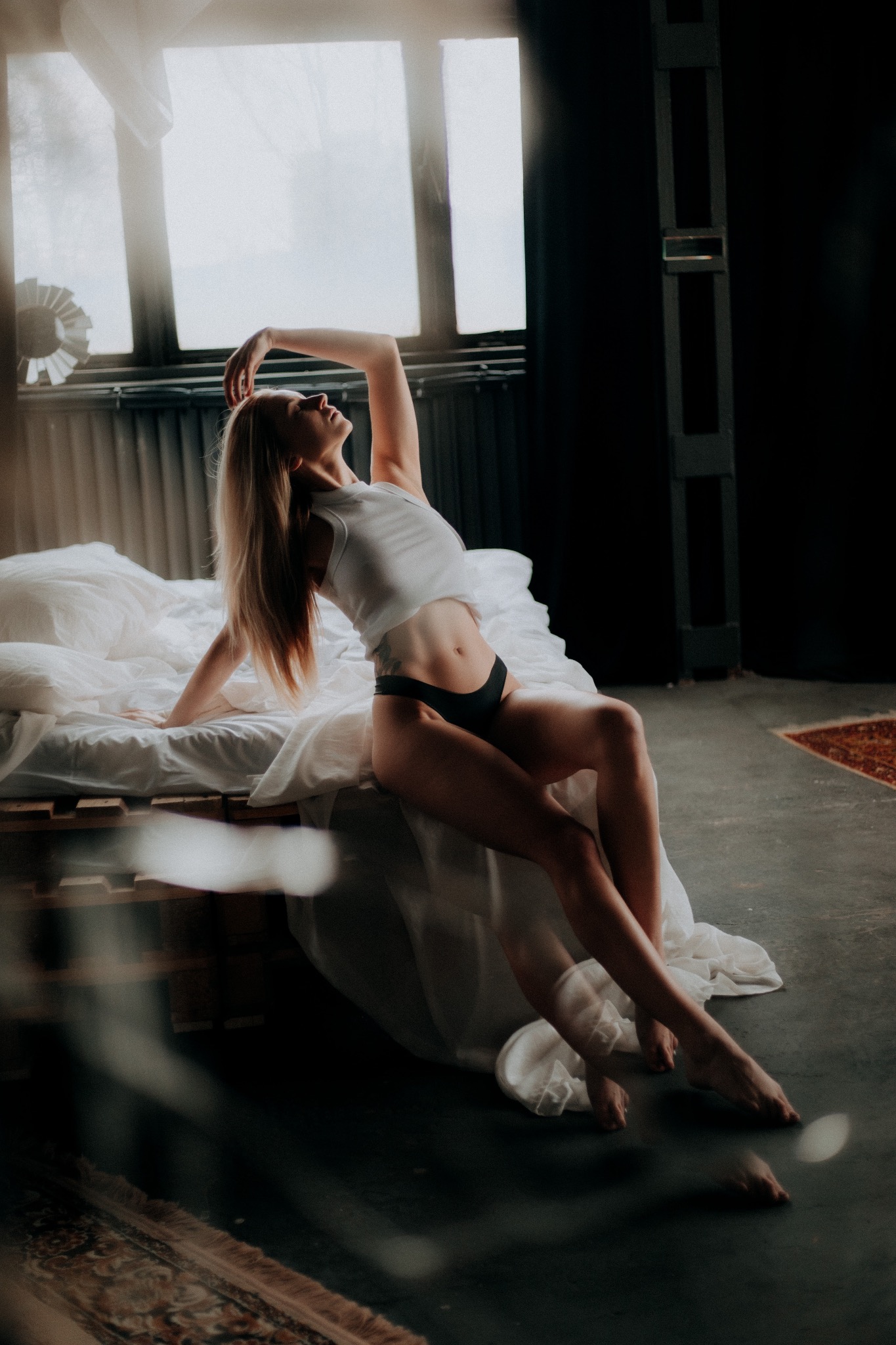 Kobieta w bieliźnie i topie pozuje na łóżku z drewnianą ramą, oświetlona przez okno w industrialnym wnętrzu z betonową podłogą i dywanem w tle.