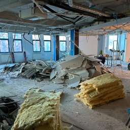 FS INVEST SPÓŁKA Z OGRANICZONĄ ODPOWIEDZIALNOŚCIĄ - Wnętrze w trakcie demontażu: sterta gruzu, odsłonięte instalacje pod sufitem, okna z widokiem na drzewa. Widoczna drabina i worki z materiałami.