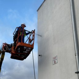 FS INVEST SPÓŁKA Z OGRANICZONĄ ODPOWIEDZIALNOŚCIĄ - Pracownik w podnośniku koszowym czyści elewację budynku myjką ciśnieniową. Widoczny fragment elewacji z tabliczką zakazu gry w piłkę.