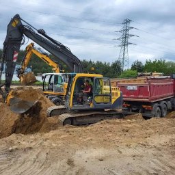 FS INVEST SPÓŁKA Z OGRANICZONĄ ODPOWIEDZIALNOŚCIĄ - Żółta koparka Volvo nabiera ziemię do czerwonej ciężarówki na placu budowy. W tle druga koparka i słup wysokiego napięcia.