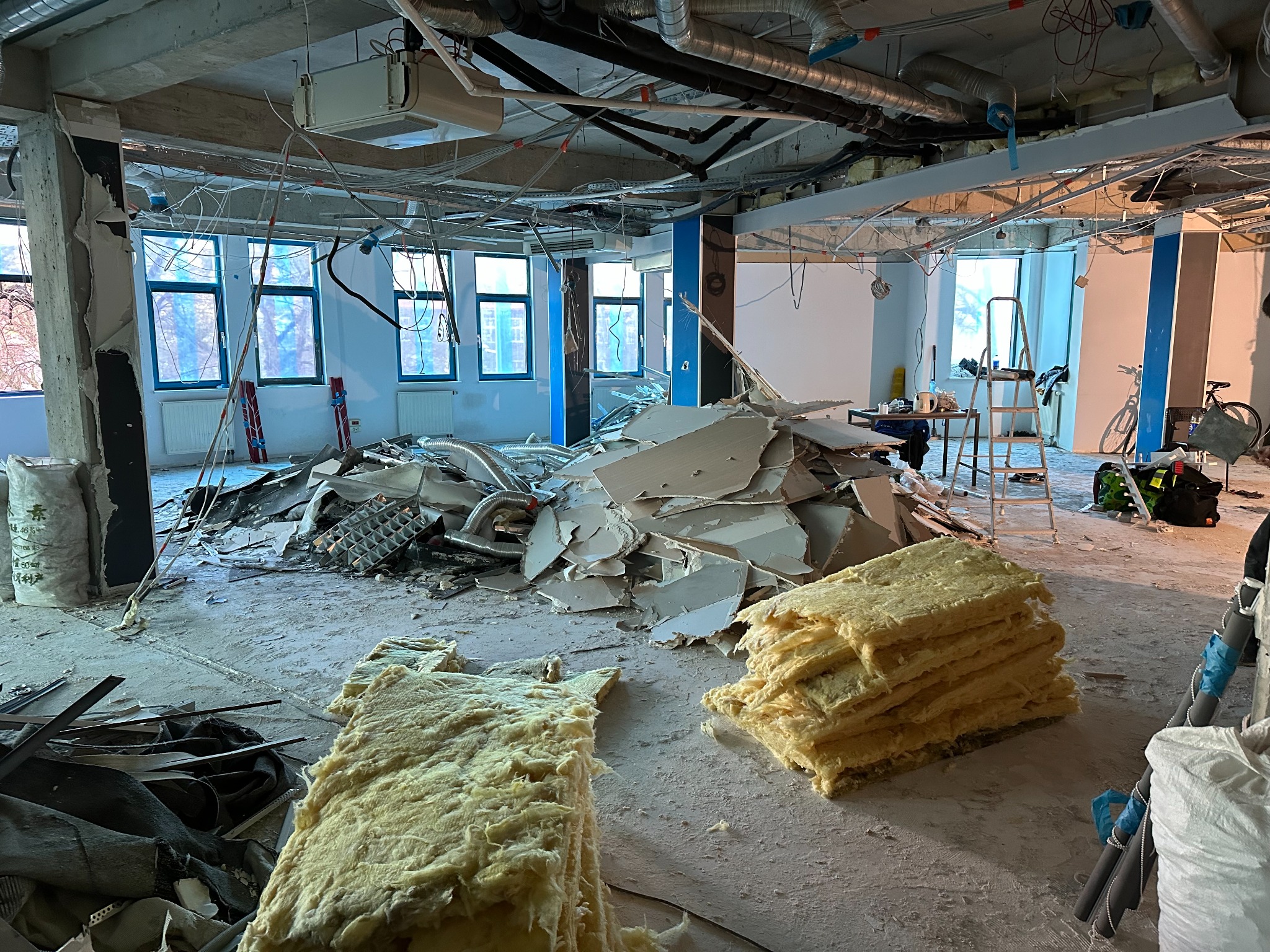 Wnętrze w trakcie demontażu: sterta gruzu, odsłonięte instalacje pod sufitem, okna z widokiem na drzewa. Widoczna drabina i worki z materiałami.