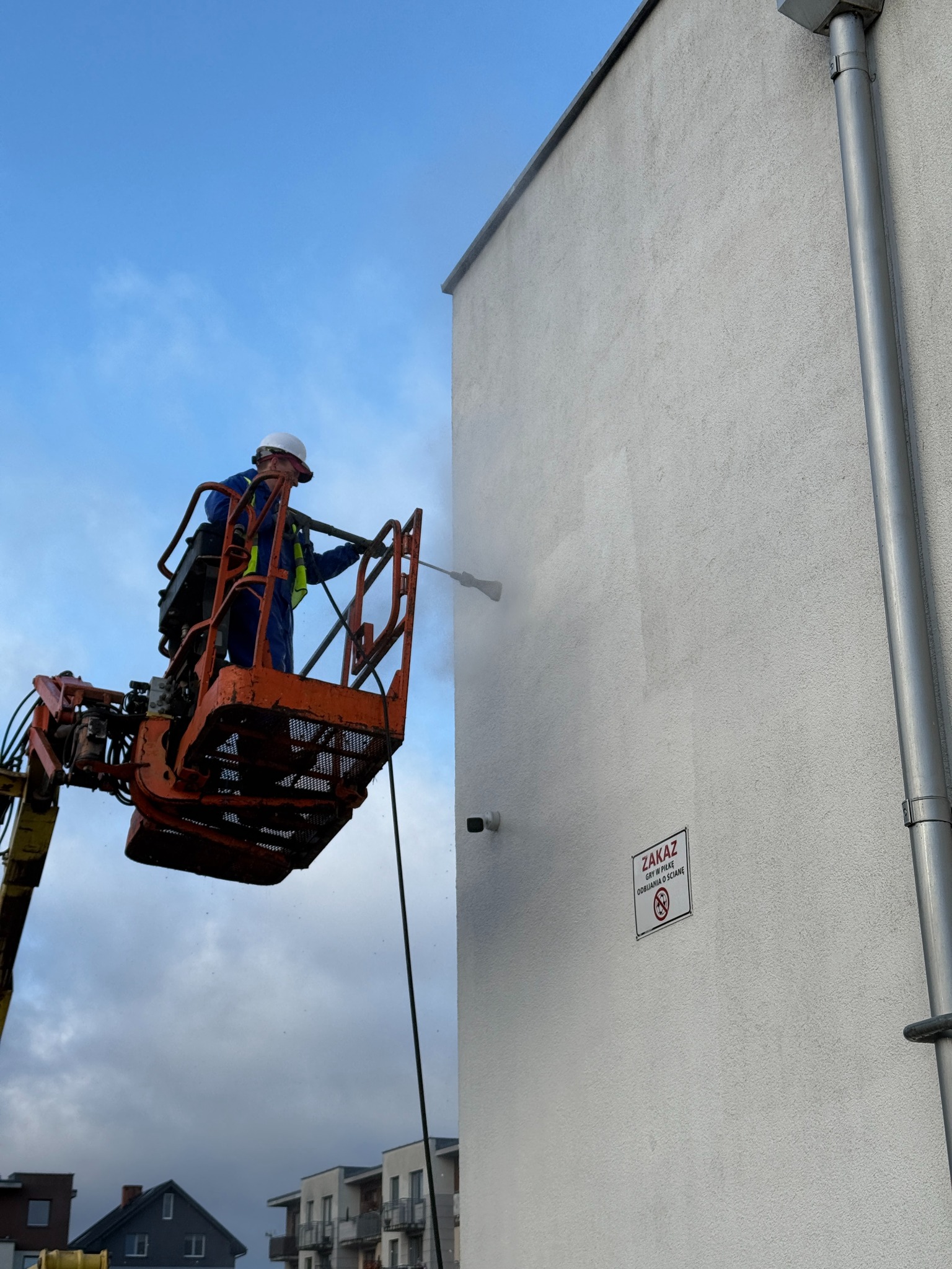 Pracownik w podnośniku koszowym czyści elewację budynku myjką ciśnieniową. Widoczny fragment elewacji z tabliczką zakazu gry w piłkę.