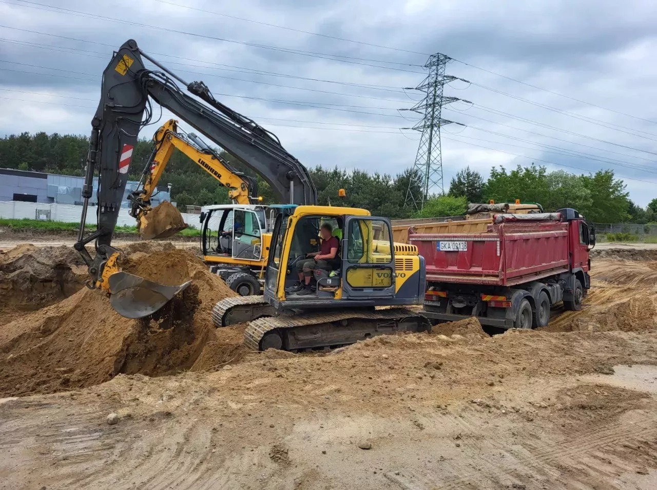Żółta koparka Volvo nabiera ziemię do czerwonej ciężarówki na placu budowy. W tle druga koparka i słup wysokiego napięcia.