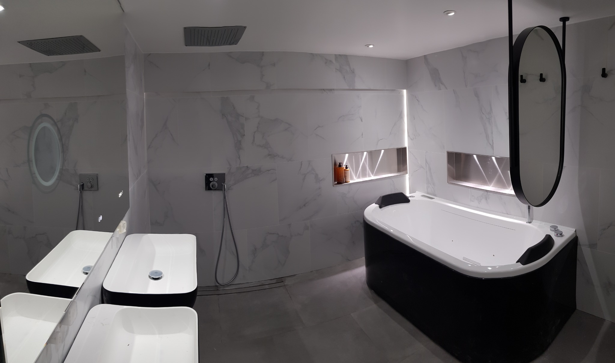 Nowoczesna łazienka z biało-czarną wanną z hydromasażem, umywalkami nablatowymi i marmurowymi płytkami ściennymi z podświetlanymi wnękami.