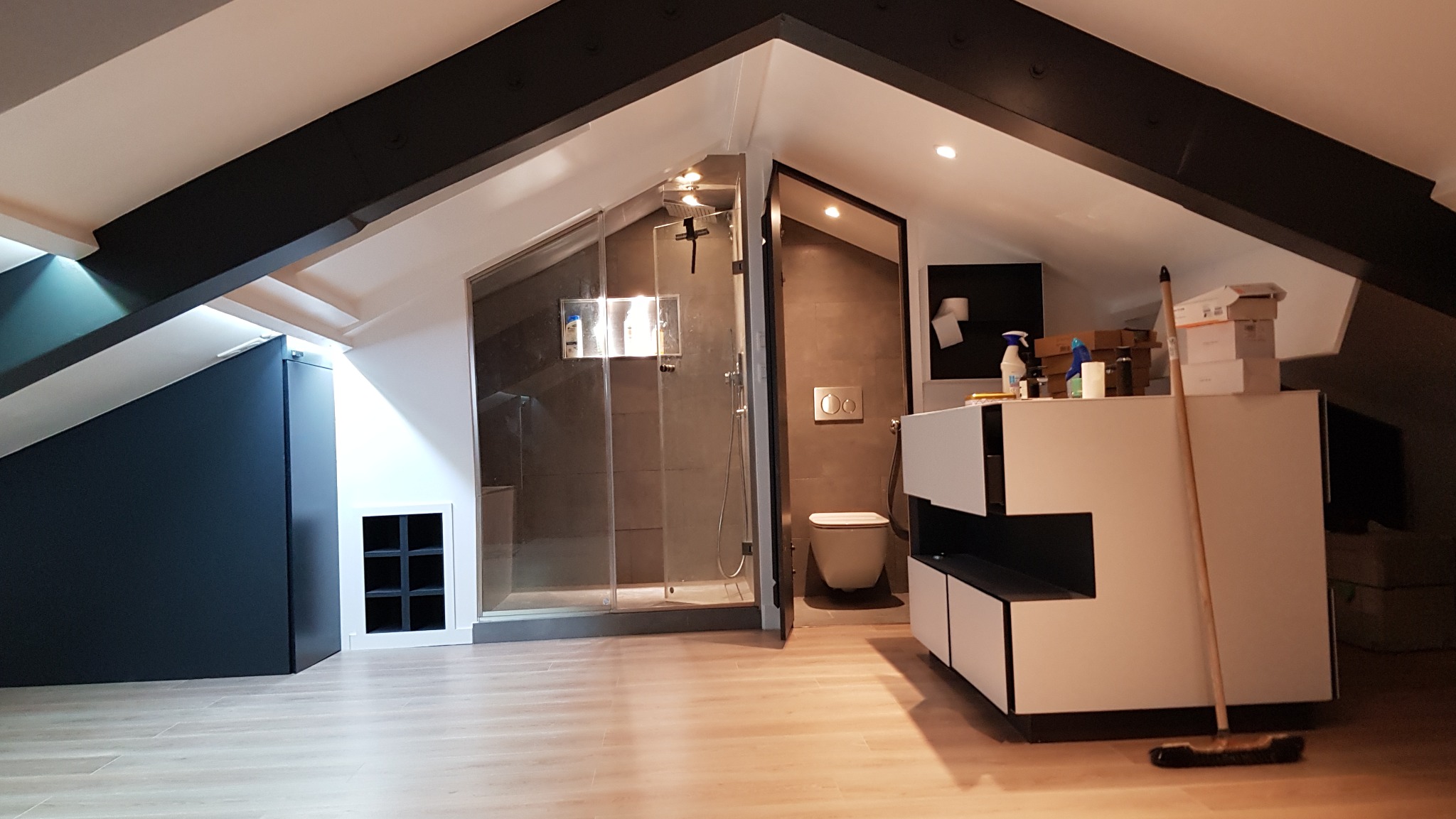 Adaptacja poddasza: łazienka z kabiną prysznicową w szarości, toaleta i umywalka z szafką, widoczny schowek w skosie dachu oraz czarne belki konstrukcyjne.