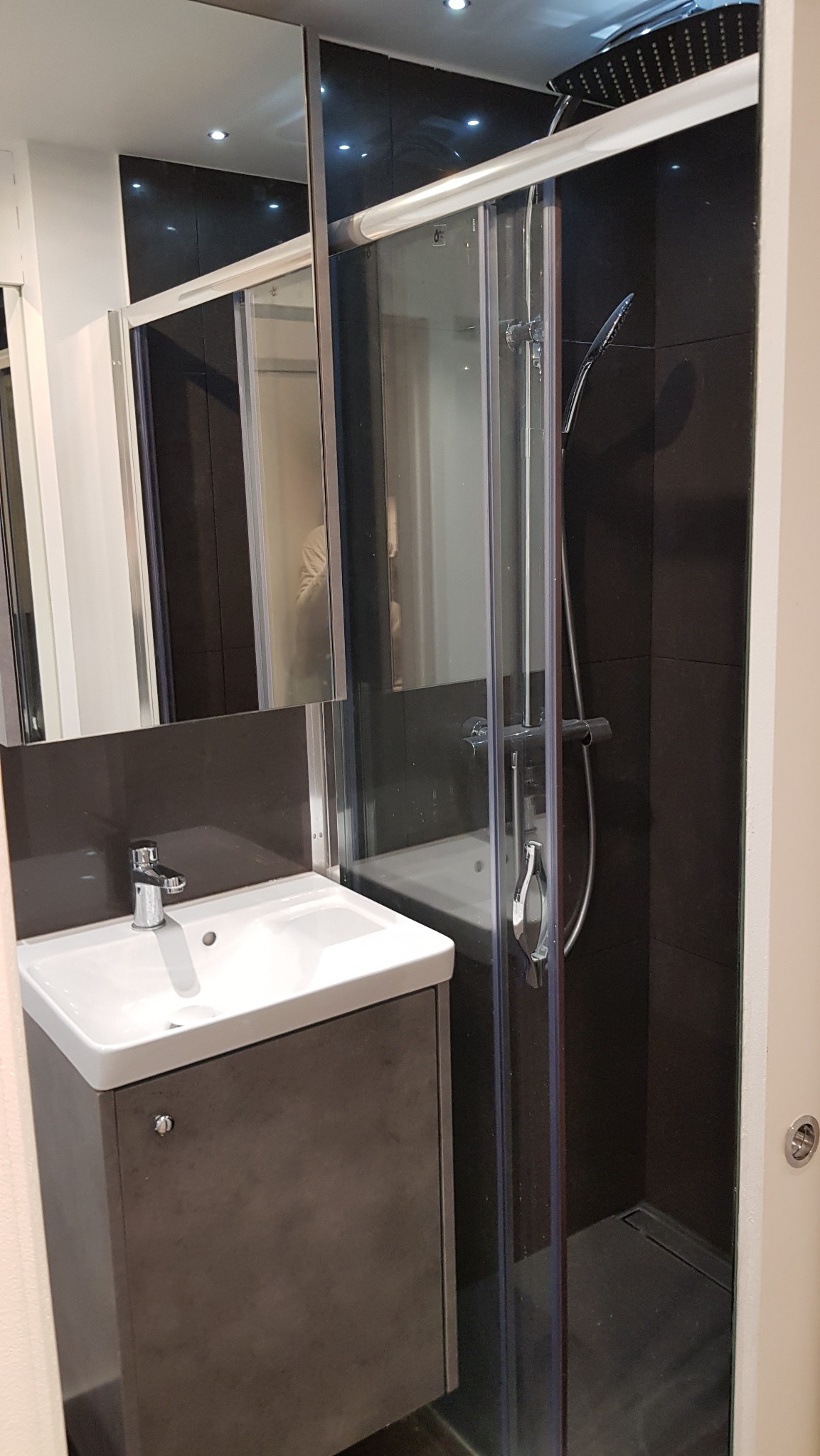 Nowoczesna łazienka z kabiną prysznicową za szklanymi drzwiami, umywalką na szafce w odcieniach szarości i czarnymi płytkami na ścianach.