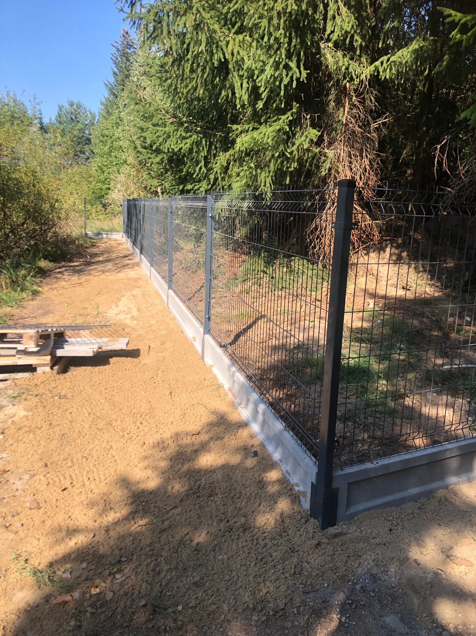 Nowo postawione, szare ogrodzenie panelowe z podmurówką betonową na nierównym, piaszczystym terenie, z widocznym lasem iglastym w tle.