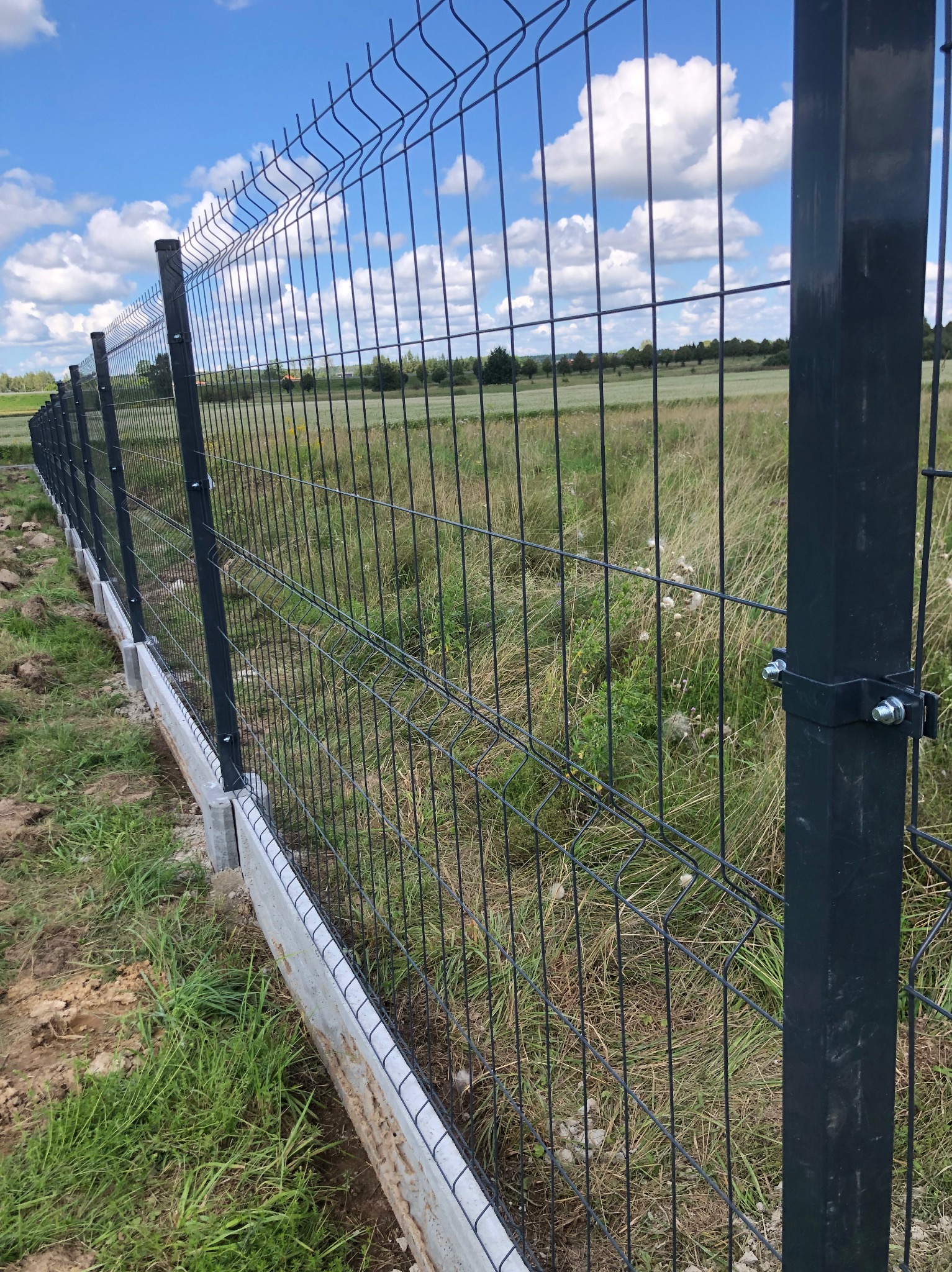Nowo zamontowane ogrodzenie panelowe z podmurówką betonową na tle zielonego pola i błękitnego nieba z chmurami.