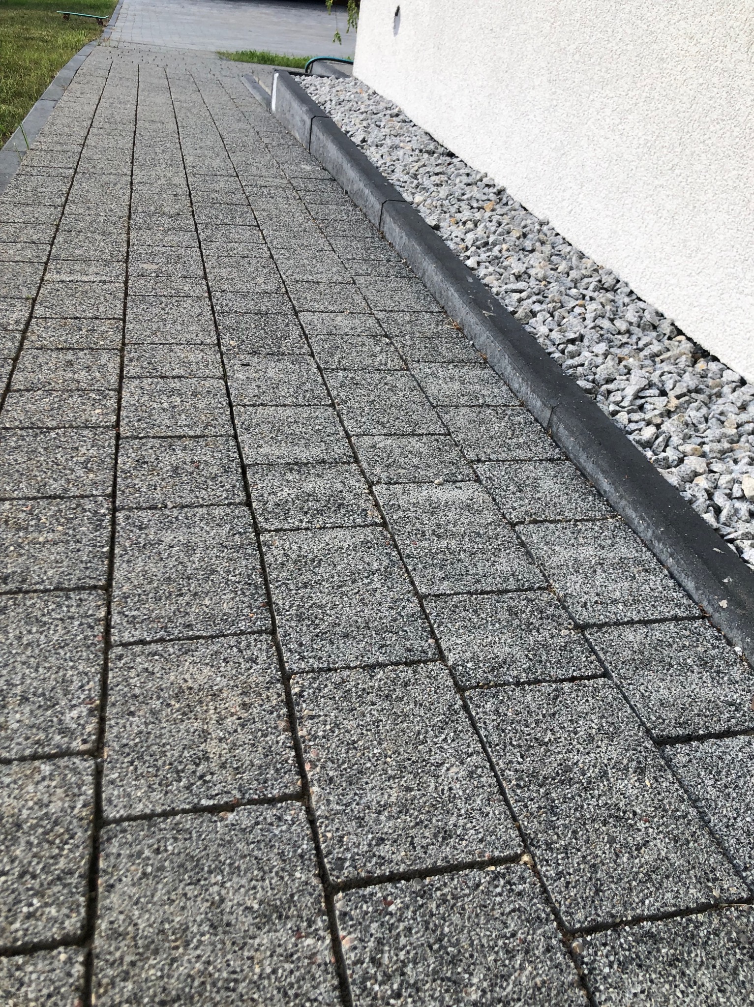 Ścieżka z szarej kostki brukowej prowadząca wzdłuż białej ściany budynku, wykończona czarnym obrzeżem i drobnym żwirem