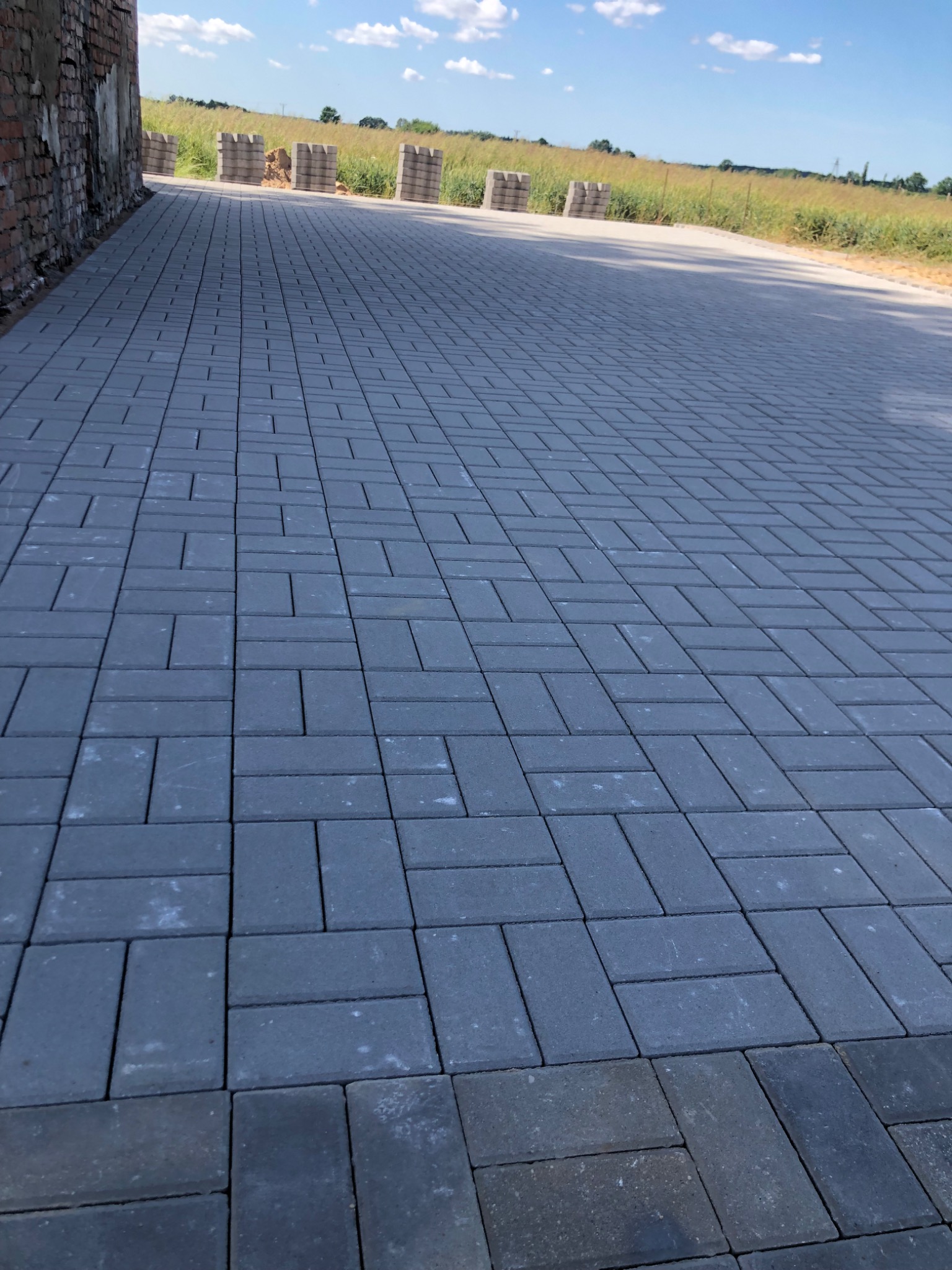 Szara kostka brukowa ułożona w regularny wzór, widok z perspektywy, w tle budynek z cegły i pole. Prezentacja gotowej nawierzchni.