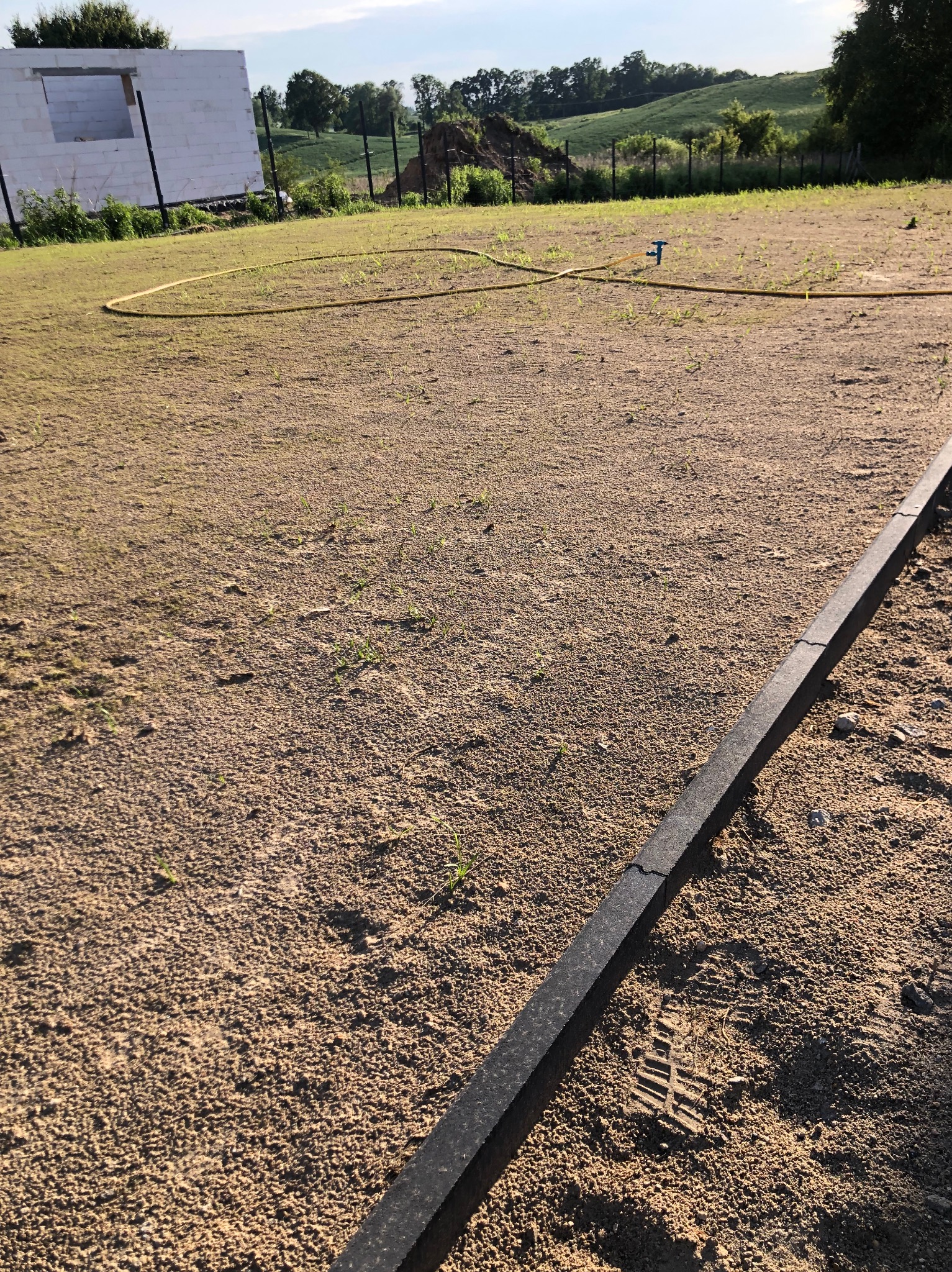 Przygotowany teren pod trawnik z obrzeżem, widoczny wąż ogrodowy i w tle budynek w stanie surowym otwartym.