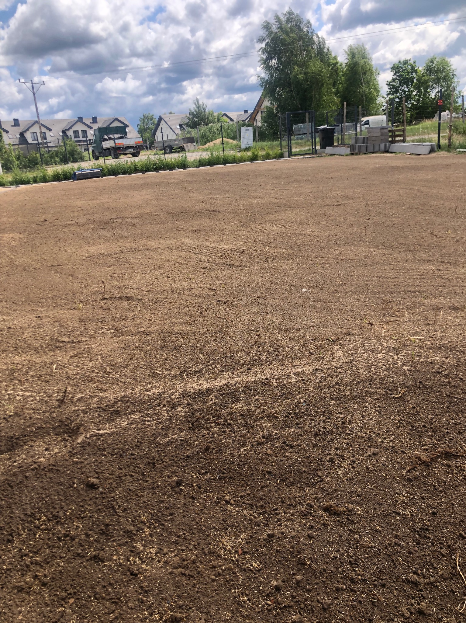 Przygotowany teren pod budowę z widoczną świeżo wyrównaną ziemią i elementami przyszłego ogrodzenia w tle, w słoneczny dzień.