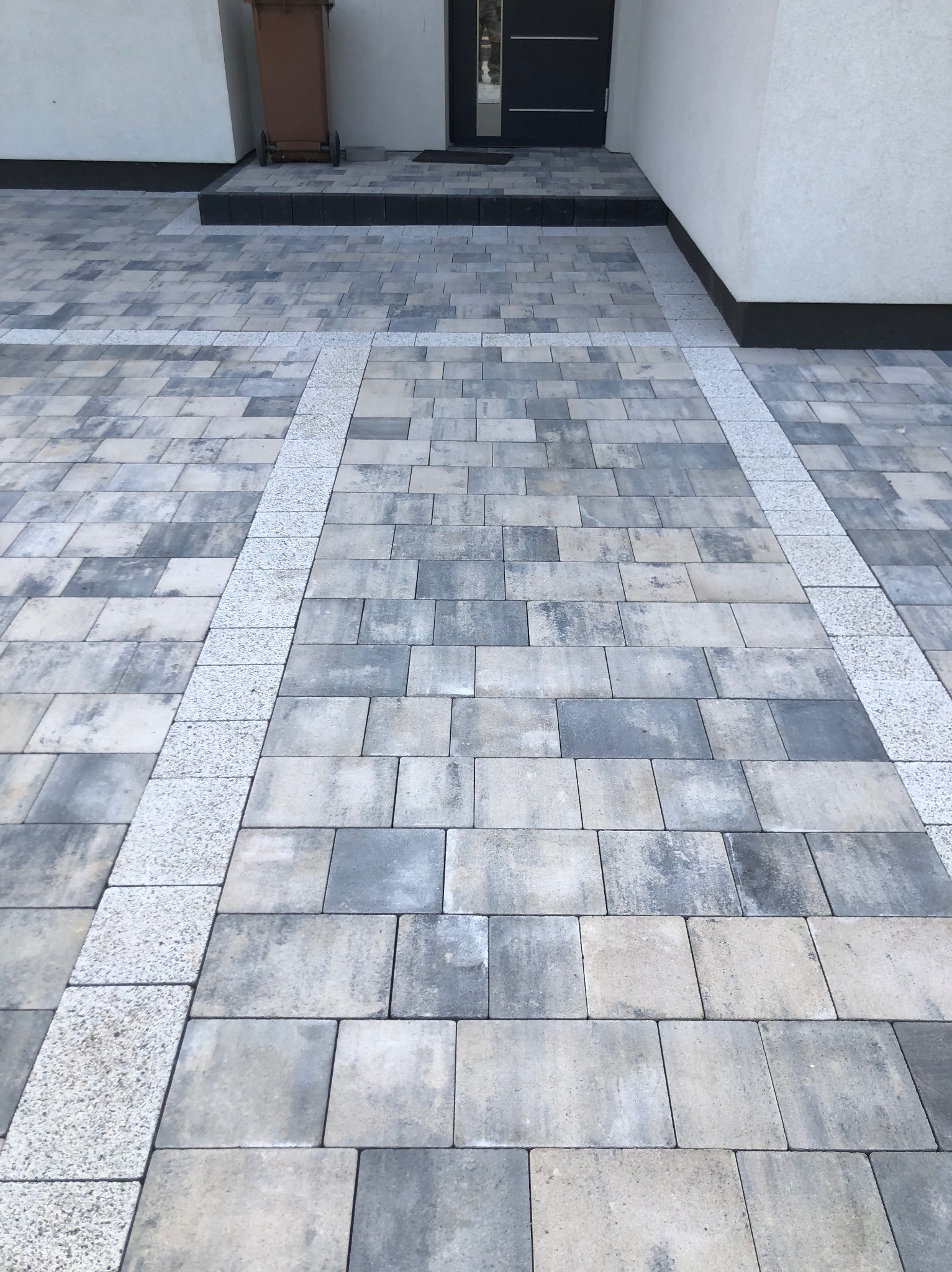 Ułożona kostka brukowa w odcieniach szarości z obramowaniem z jasnej kostki granitowej. Widok na podjazd przed domem z ciemnymi drzwiami wejściowymi.