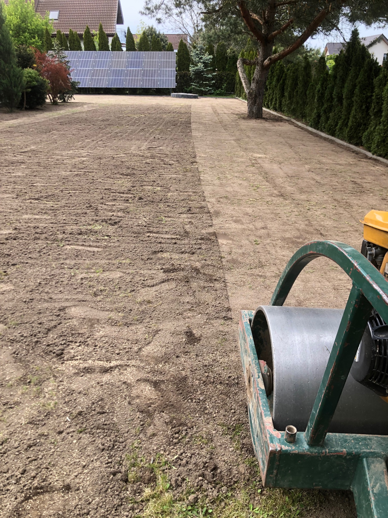 Przygotowanie gruntu pod trawnik walcem ogrodowym. Częściowo wyrównana ziemia, widoczne ślady po walcu. W tle panele słoneczne i drzewa.