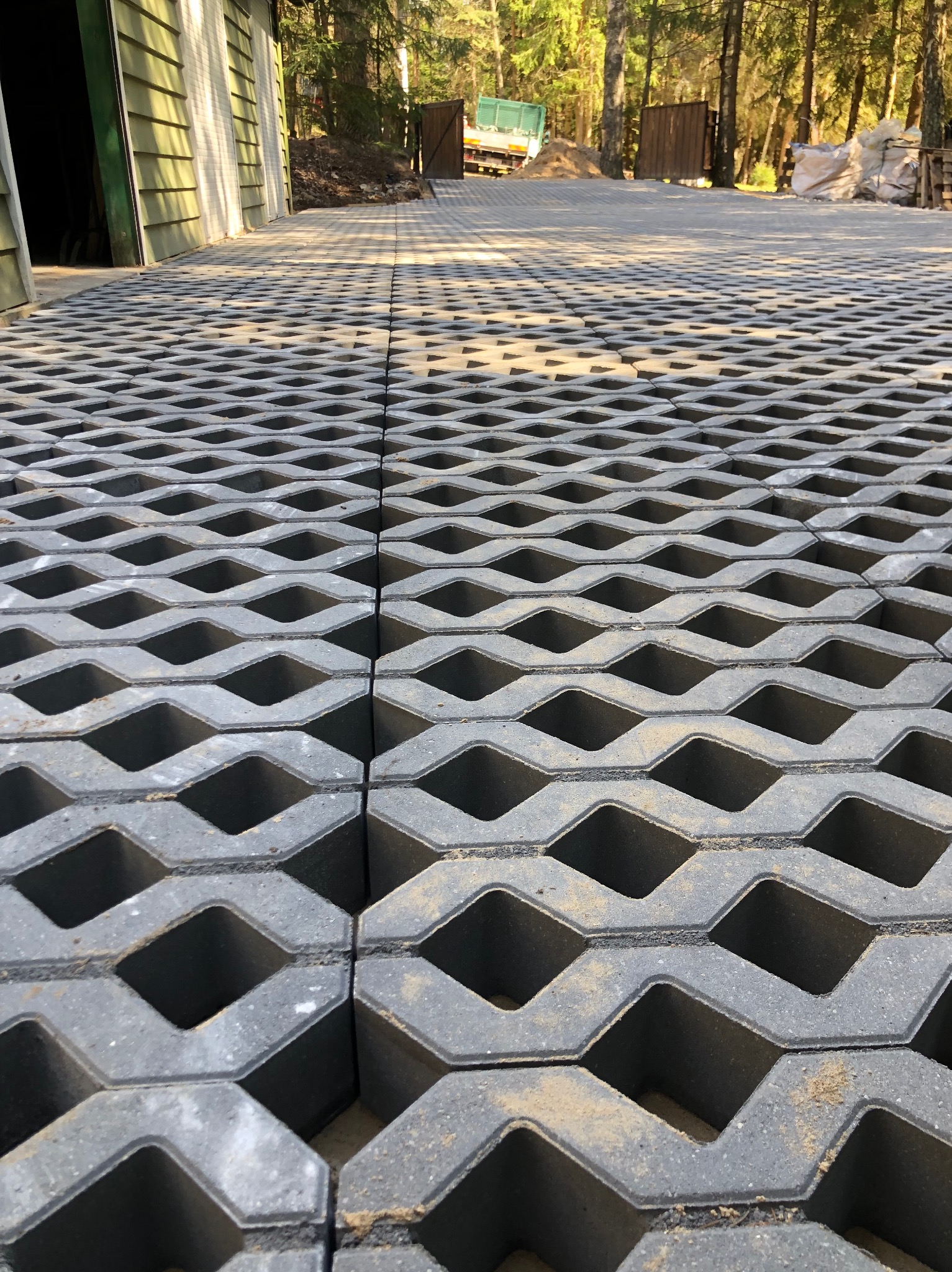 Szara, ażurowa kostka brukowa ułożona na podjeździe, z widocznym budynkiem w tle i elementami zieleni. Perspektywa z bliska podkreśla strukturę i wzór kostki.