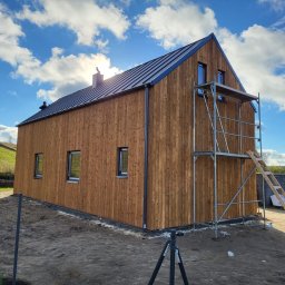 FIRMA HANDLOWO USŁUGOWA DAWID KOWALCZYK - Nowoczesny dom z drewnianą elewacją w trakcie budowy, z widocznym rusztowaniem i drabiną, na tle błękitnego nieba z chmurami.