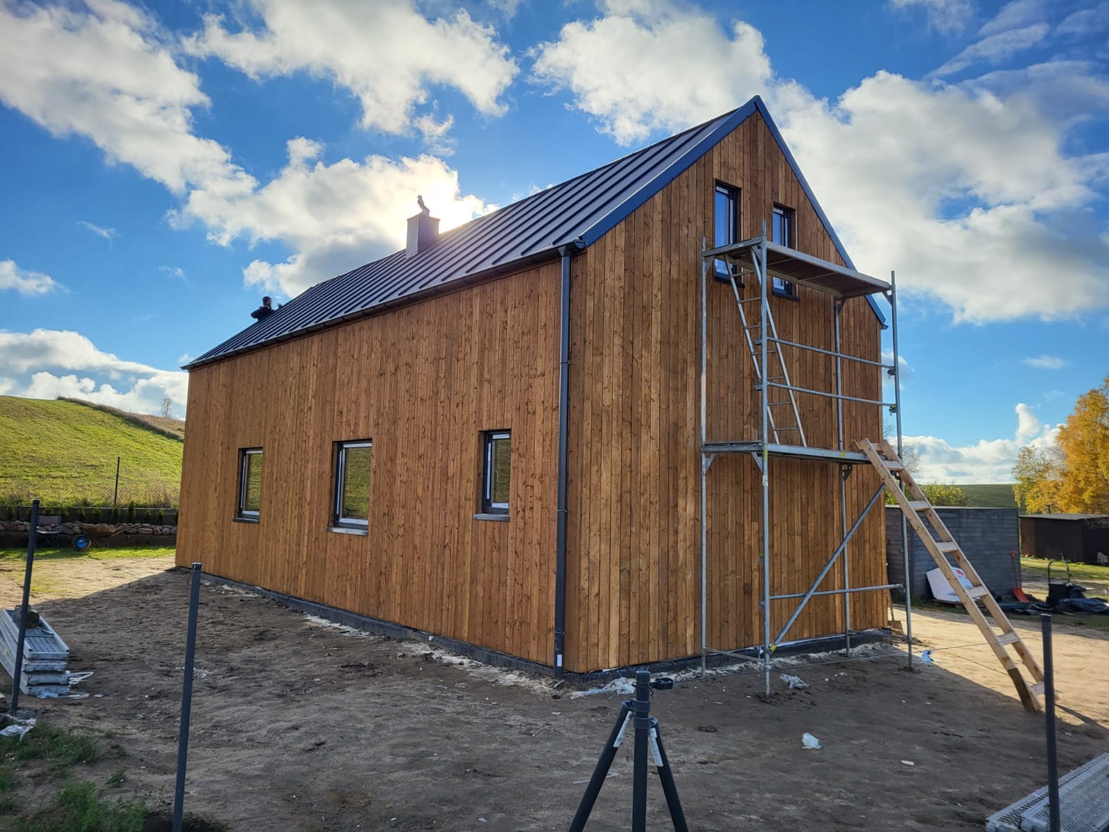 Nowoczesny dom z drewnianą elewacją w trakcie budowy, z widocznym rusztowaniem i drabiną, na tle błękitnego nieba z chmurami.