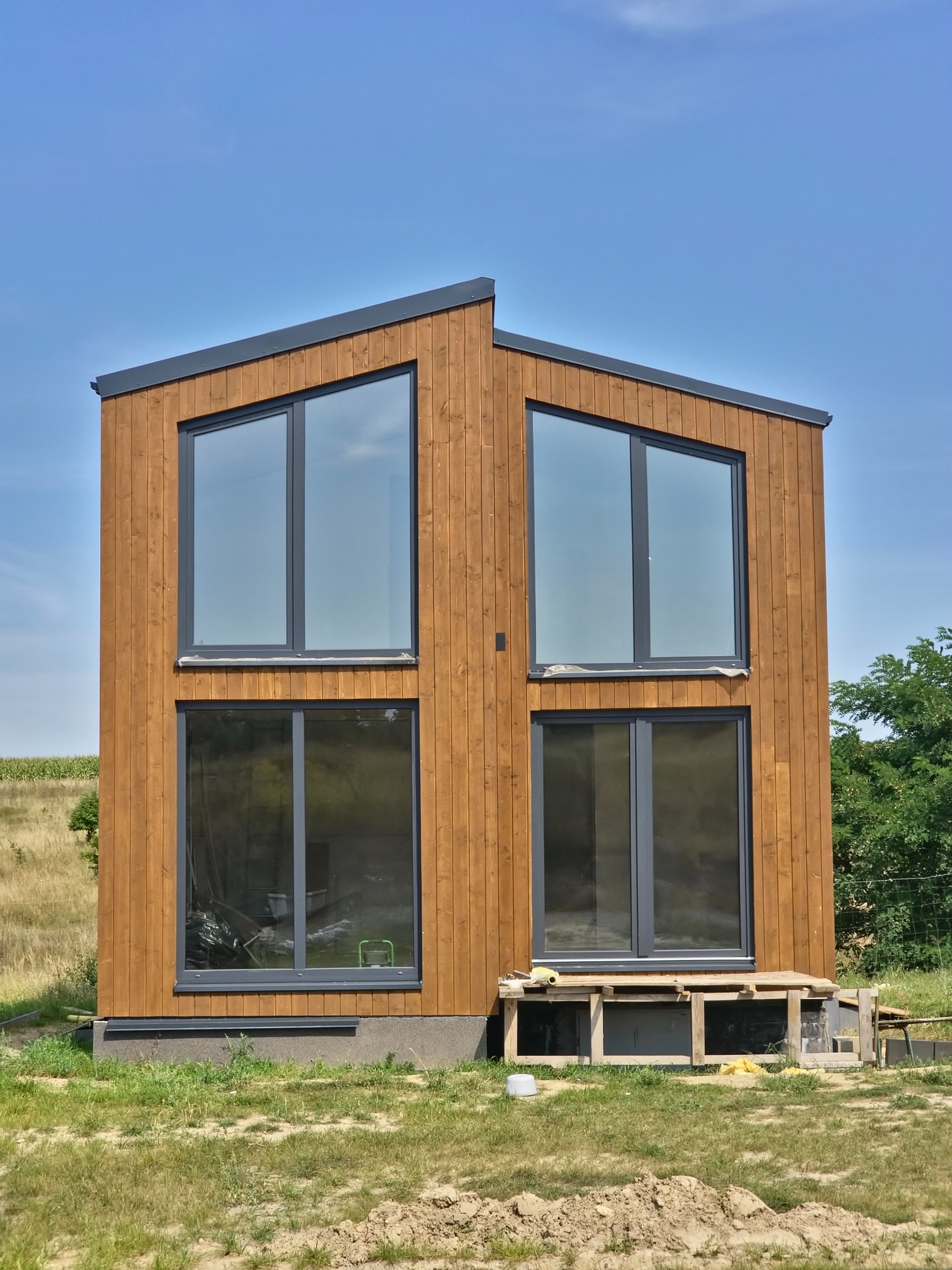 Nowoczesny, dwukondygnacyjny budynek z drewnianą elewacją i dużymi oknami, w trakcie budowy, z widocznymi tymczasowymi podporami i elementami placu budowy.