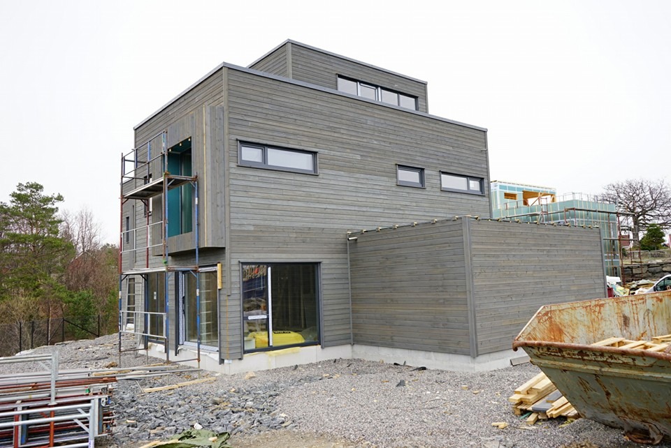 Surowy, nowoczesny dom z szarego drewna w trakcie budowy, z rusztowaniami, widocznymi elementami konstrukcyjnymi i kontenerem na gruz w tle.