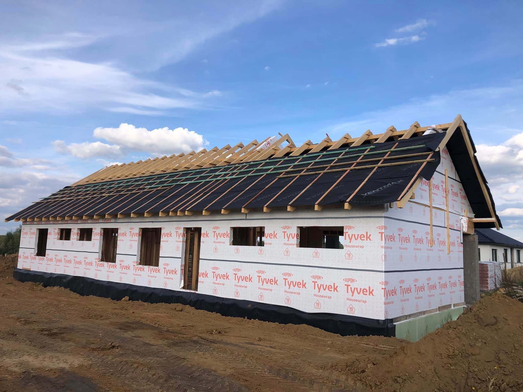 Konstrukcja domu szkieletowego w trakcie budowy: widoczne ściany obłożone membraną Tyvek, drewniany szkielet dachu z czarną membraną dachową, ziemia wokół budynku.