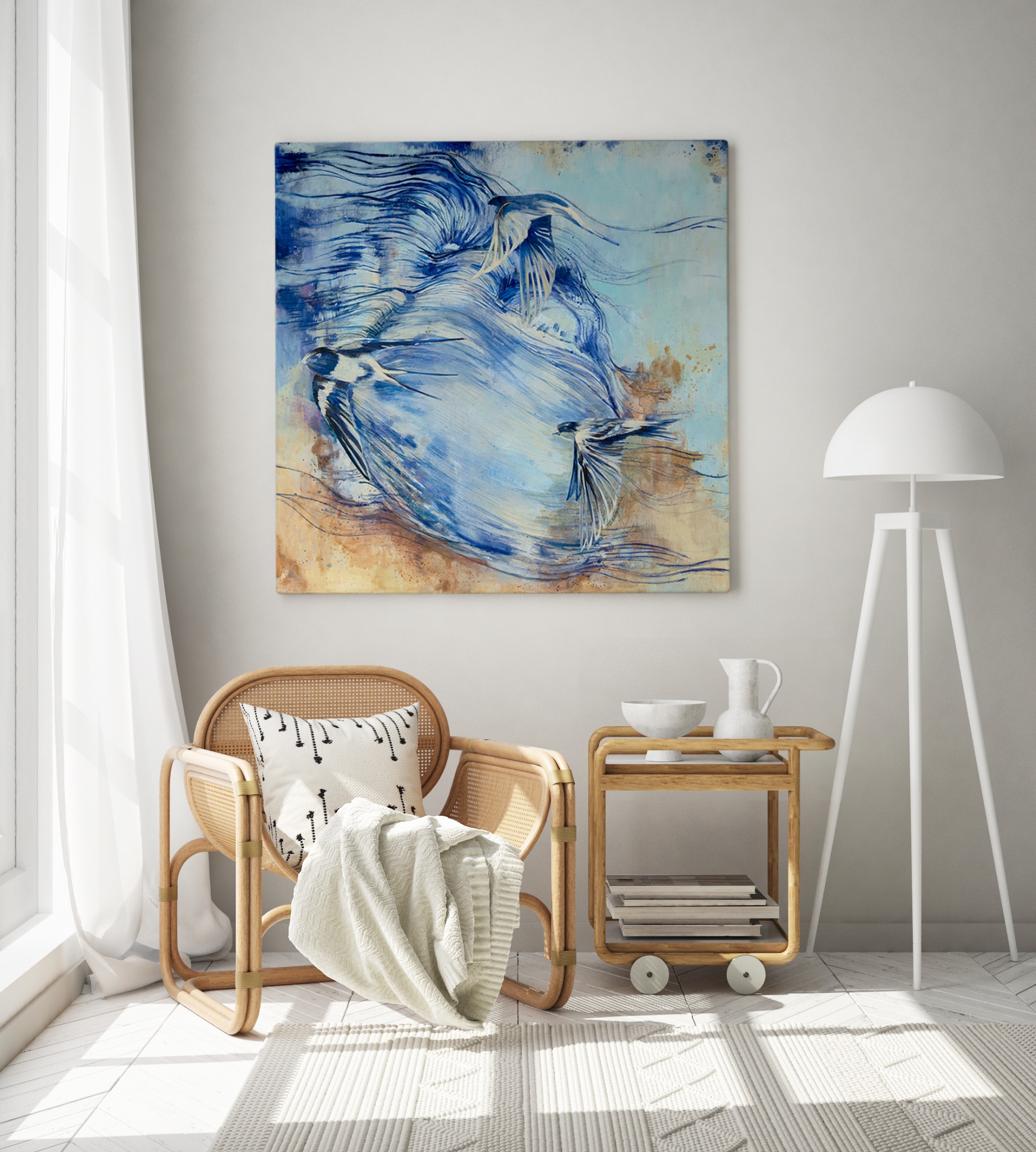 Abstrakcyjny obraz w odcieniach błękitu i beżu z motywem lecących ptaków, wiszący nad wiklinowym fotelem z poduszką i pledem w jasnym, minimalistycznym wnętrzu z lampą podłogową.