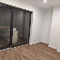 Zander Oleksandr Prishchenko - Jasne, świeżo pomalowane wnętrze z panelami podłogowymi, z widocznymi elementami przygotowanymi do dalszych prac przez duże okno balkonowe.