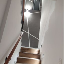 Zander Oleksandr Prishchenko - Klatka schodowa z drewnopodobnymi stopniami, białą balustradą i szarą ścianą z geometrycznym wzorem z białych linii, oświetlona małymi punktowymi lampami.