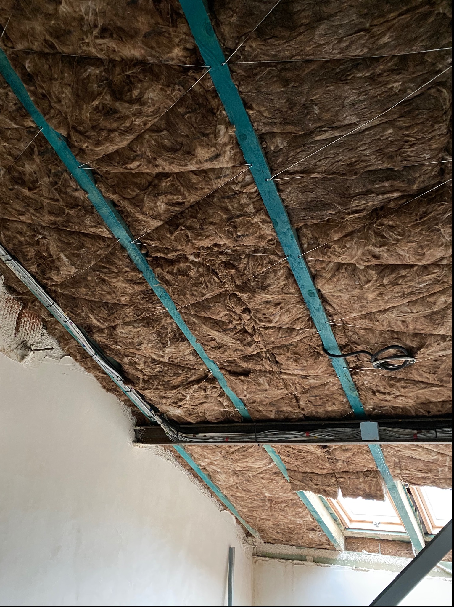 Izolacja z wełny mineralnej zamontowana na drewnianych belkach w poddaszu, zabezpieczona sznurkiem, widoczne przewody elektryczne i okno dachowe.