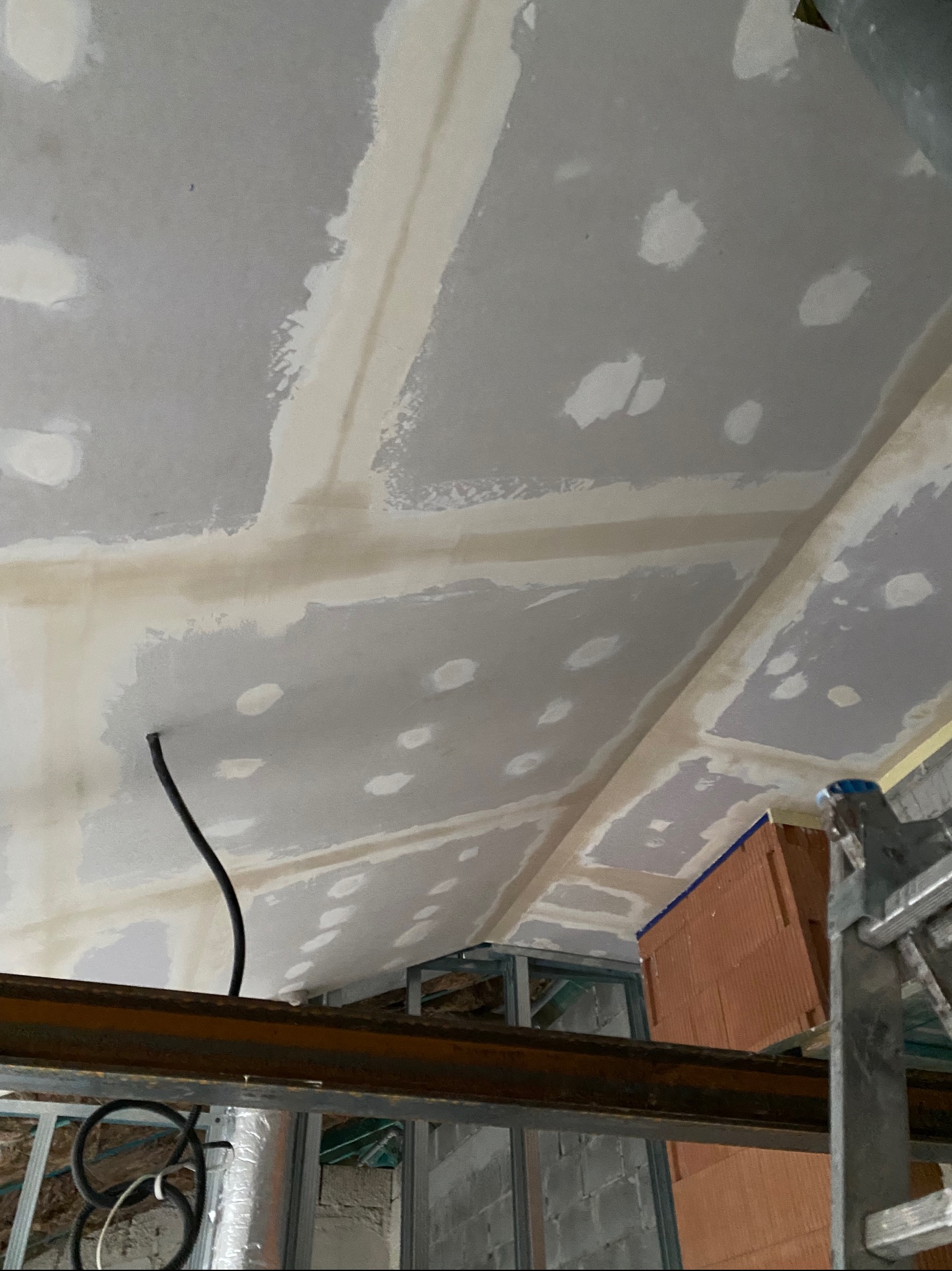 Sufit poddasza w trakcie szpachlowania, widoczne łączenia płyt gipsowo-kartonowych i metalowa konstrukcja nośna, z fragmentem czarnego przewodu elektrycznego.