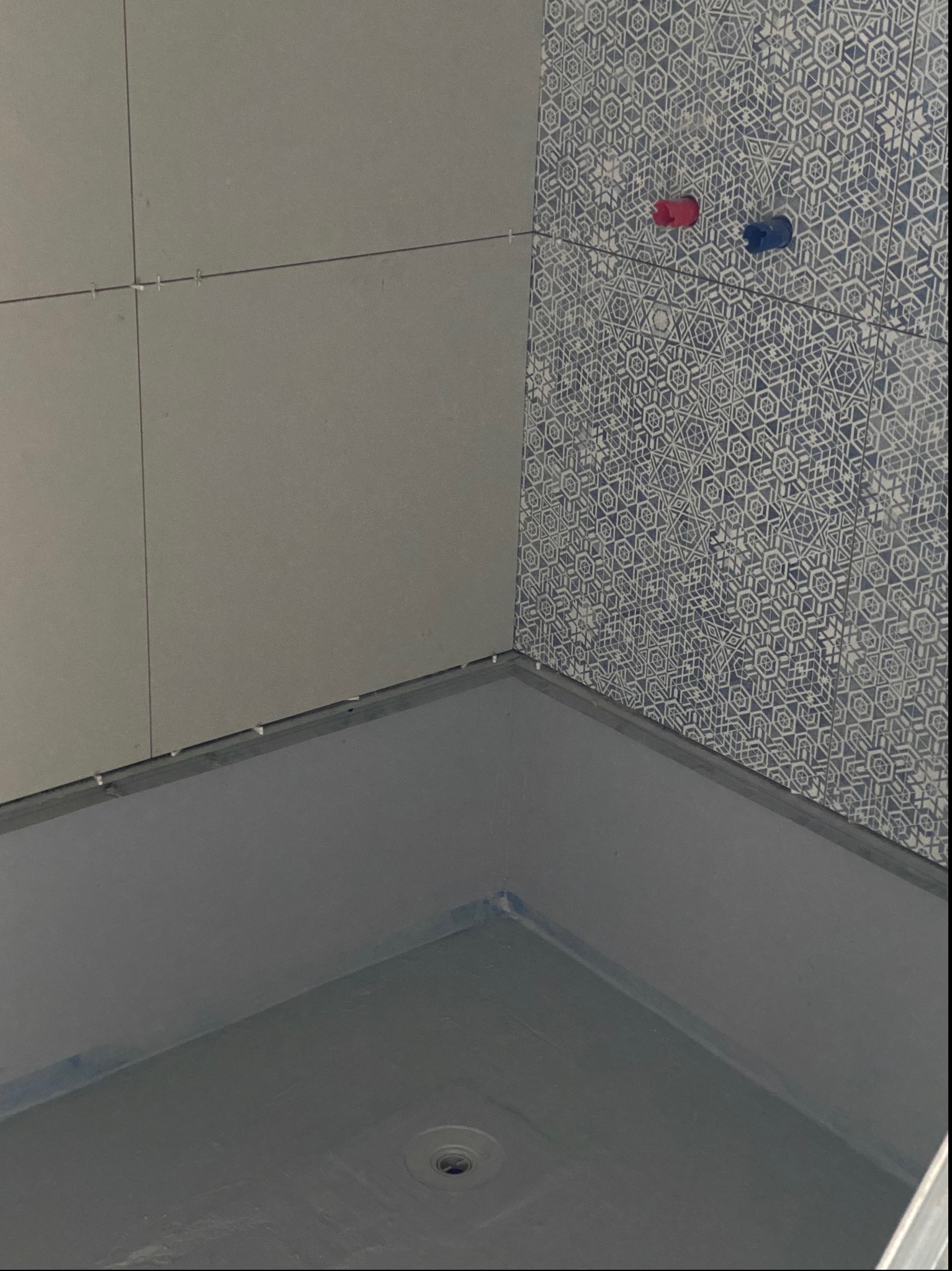 Przygotowana do dalszych prac strefa prysznicowa z uszczelnioną podłogą i częściowo ułożonymi płytkami ściennymi, widoczne krzyżyki dystansowe i rury doprowadzające wodę.