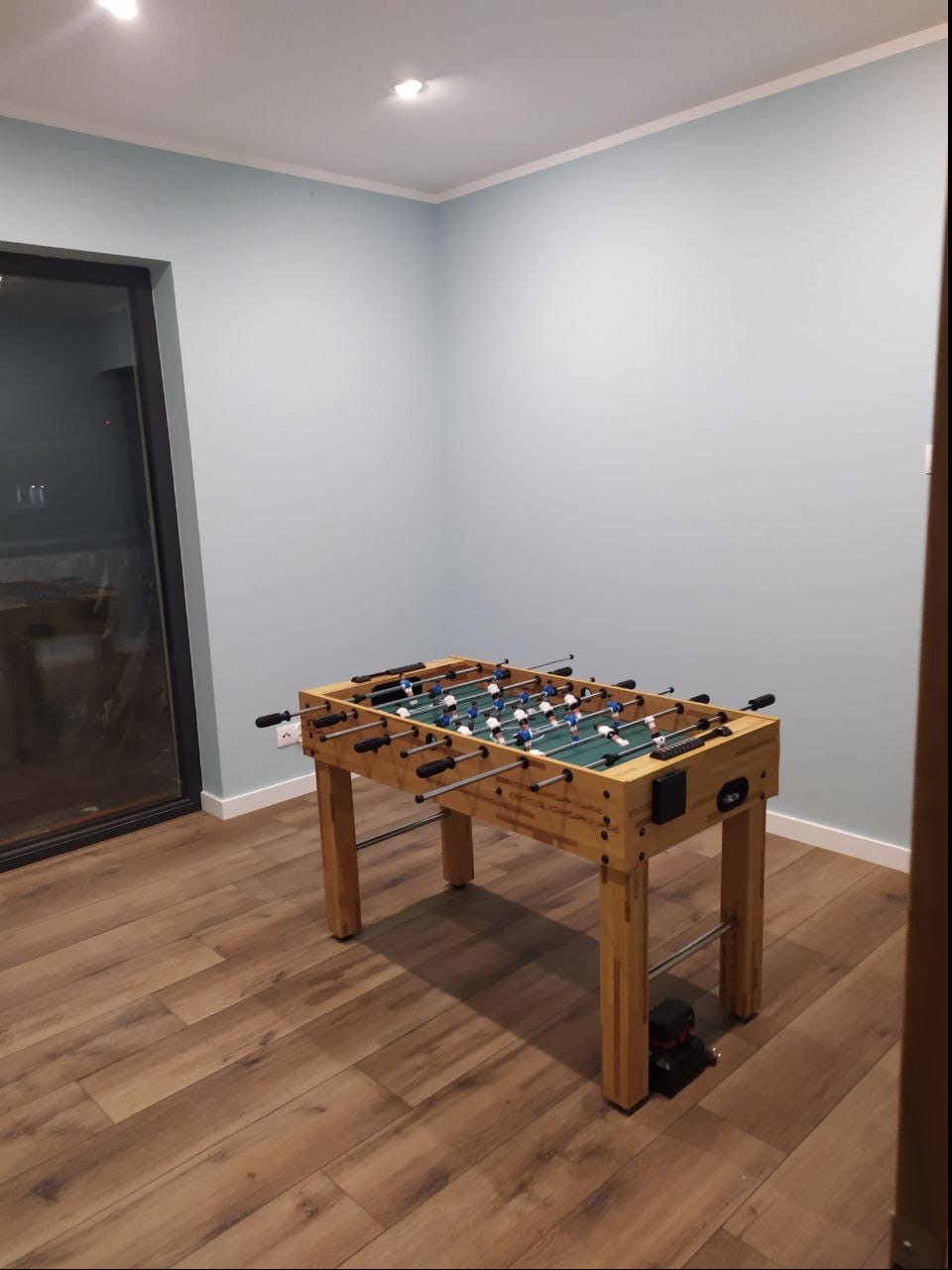 Pokój z jasnymi ścianami i drewnianą podłogą, wyposażony w stół do gry w piłkarzyki z drewnianą obudową i czarnymi uchwytami, oświetlony przez wbudowane lampy sufitowe.