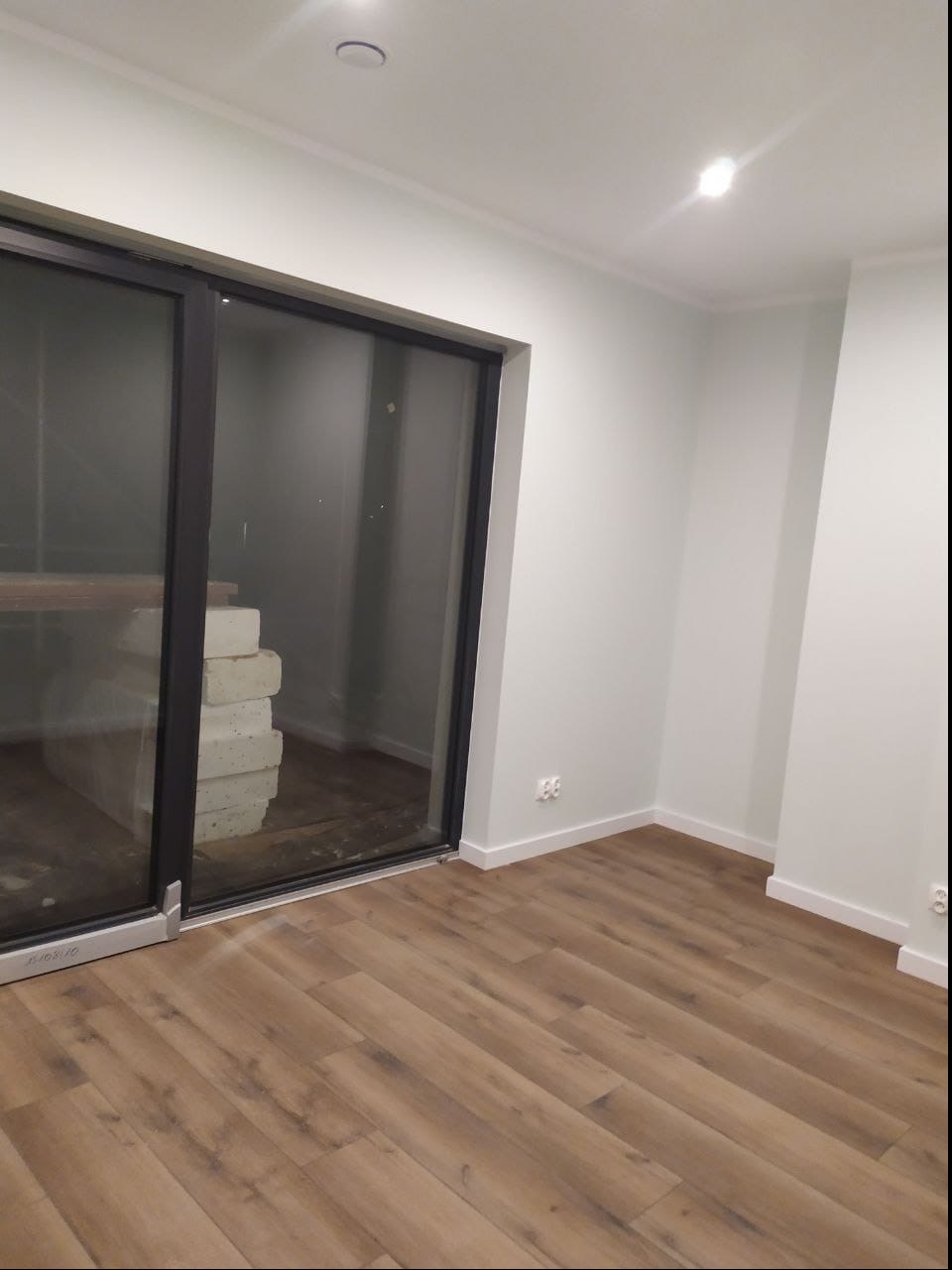 Jasne, świeżo pomalowane wnętrze z panelami podłogowymi, z widocznymi elementami przygotowanymi do dalszych prac przez duże okno balkonowe.