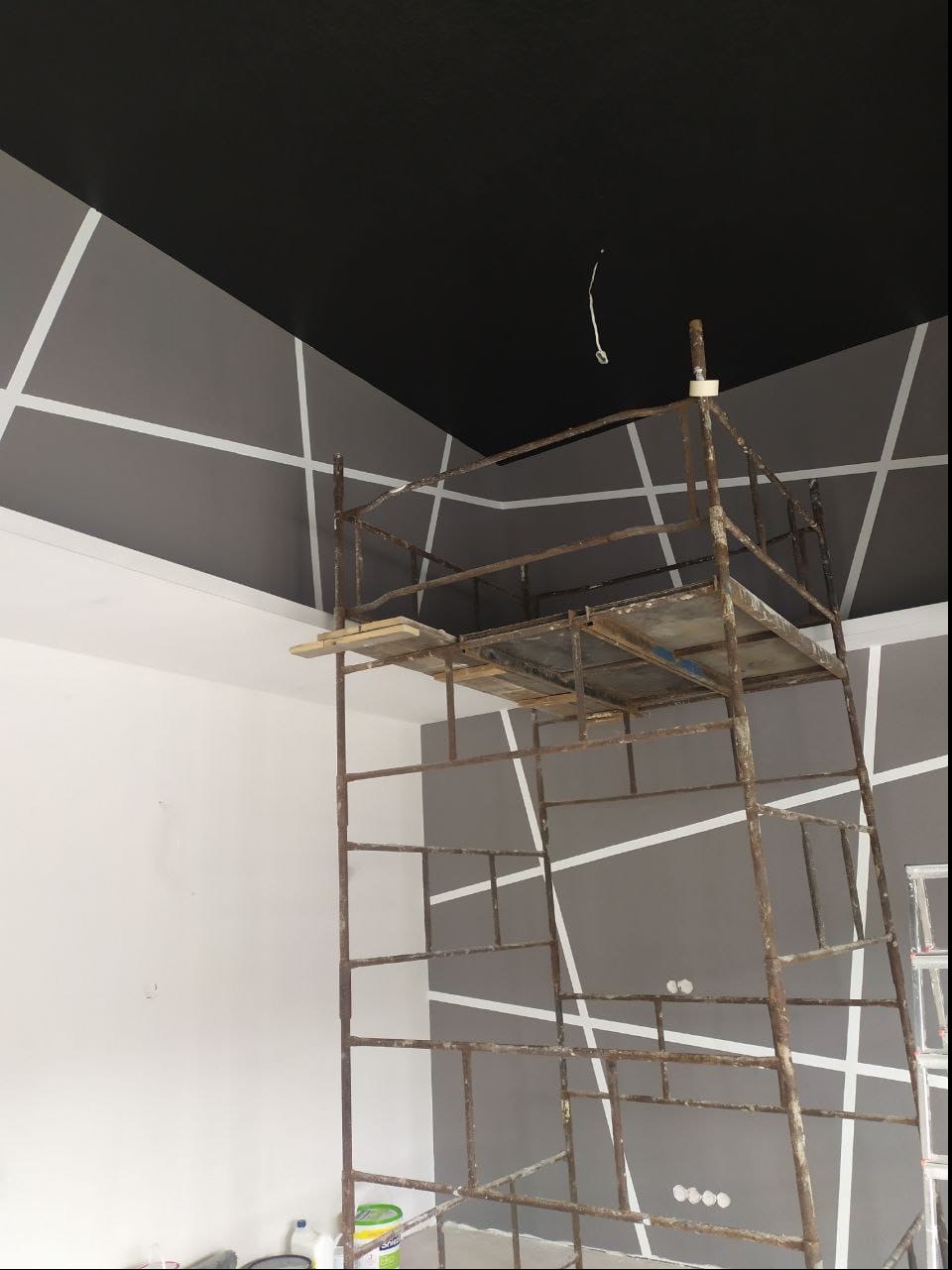 Pokój w trakcie malowania z czarnym sufitem i szarą ścianą z białymi, ukośnymi pasami, widoczne rusztowanie malarskie.