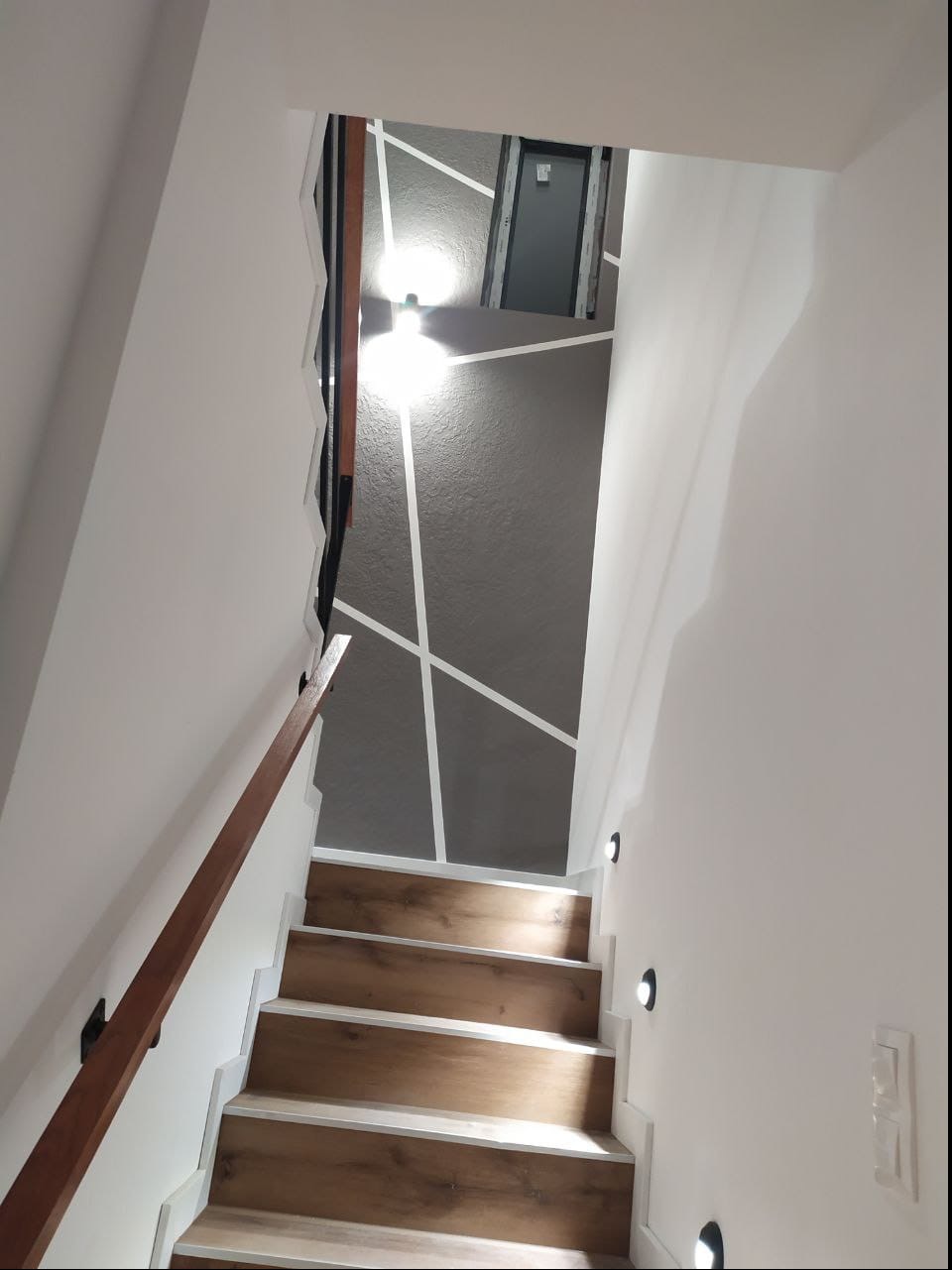 Klatka schodowa z drewnopodobnymi stopniami, białą balustradą i szarą ścianą z geometrycznym wzorem z białych linii, oświetlona małymi punktowymi lampami.