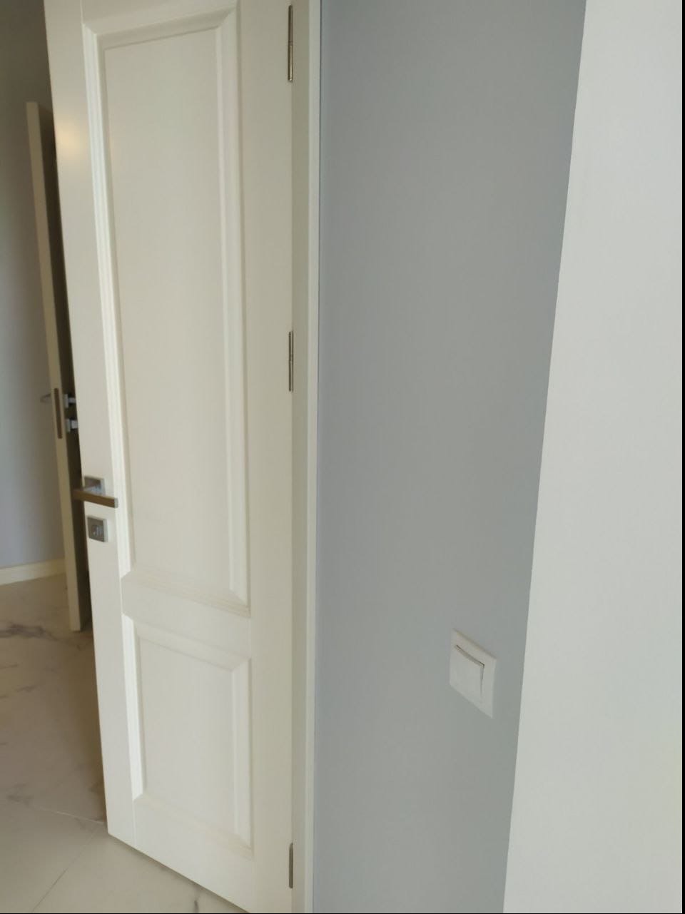 Otwarta, biała, dwuskrzydłowa drzwi wewnętrzne z srebrną klamką, na tle szarej ściany z zamontowanym włącznikiem światła.