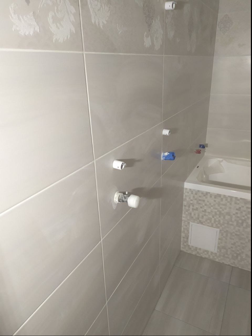 Ściana łazienki wyłożona jasnymi, prostokątnymi płytkami z delikatnym, kwiatowym wzorem u góry, widoczne białe zawory i niebieska taśma zabezpieczająca, obok fragment obudowy wanny z mozaiką.