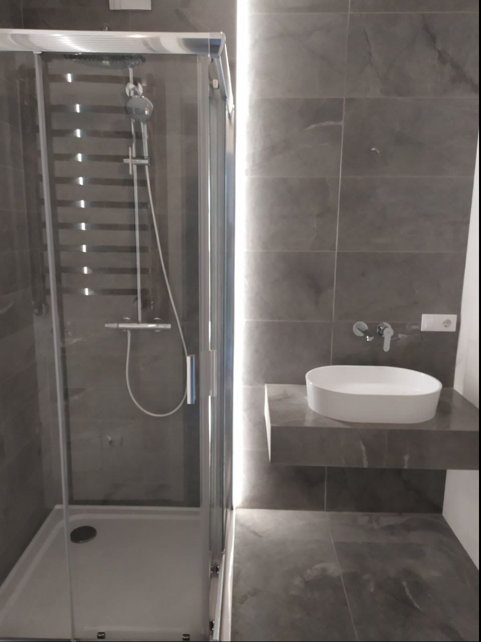 Nowoczesna łazienka z kabiną prysznicową z przezroczystego szkła, szarą armaturą i umywalką nablatową na kamiennym blacie, oświetlona pionowym paskiem LED.