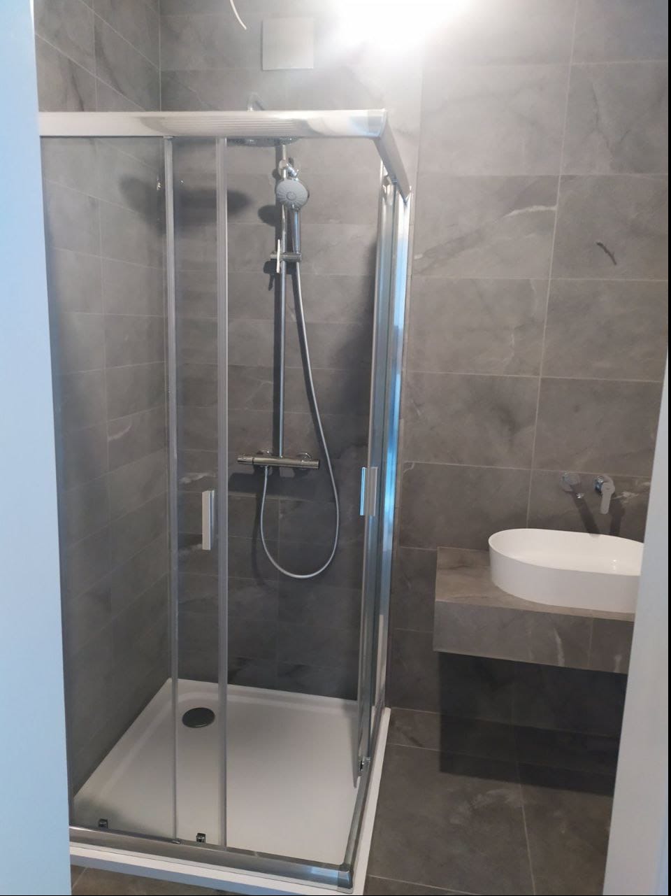 Nowoczesna kabina prysznicowa z przesuwnymi drzwiami i biały umywalka nablatowa na marmurowym blacie, szare płytki na ścianach.