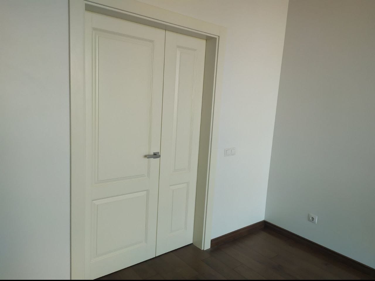 Zamknięte, białe dwuskrzydłowe drzwi z srebrną klamką, w jasnym pomieszczeniu z drewnianą podłogą i gładkimi, pomalowanymi na biało ścianami.