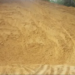Góra jasnego piasku z wyraźnymi śladami opon pojazdu budowlanego u podstawy, częściowo zacieniona u góry, z fragmentem zieleni w tle.